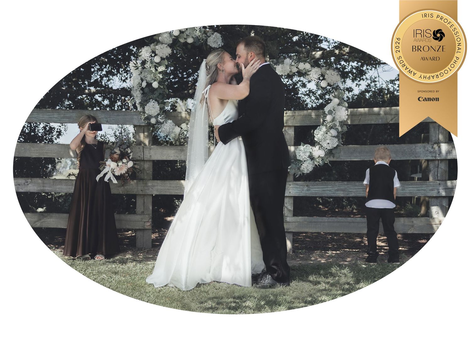 A bride and groom share a kiss at an outdoor wedding ceremony by a rustic wooden fence with a floral arch backdrop, by Charly Baty.