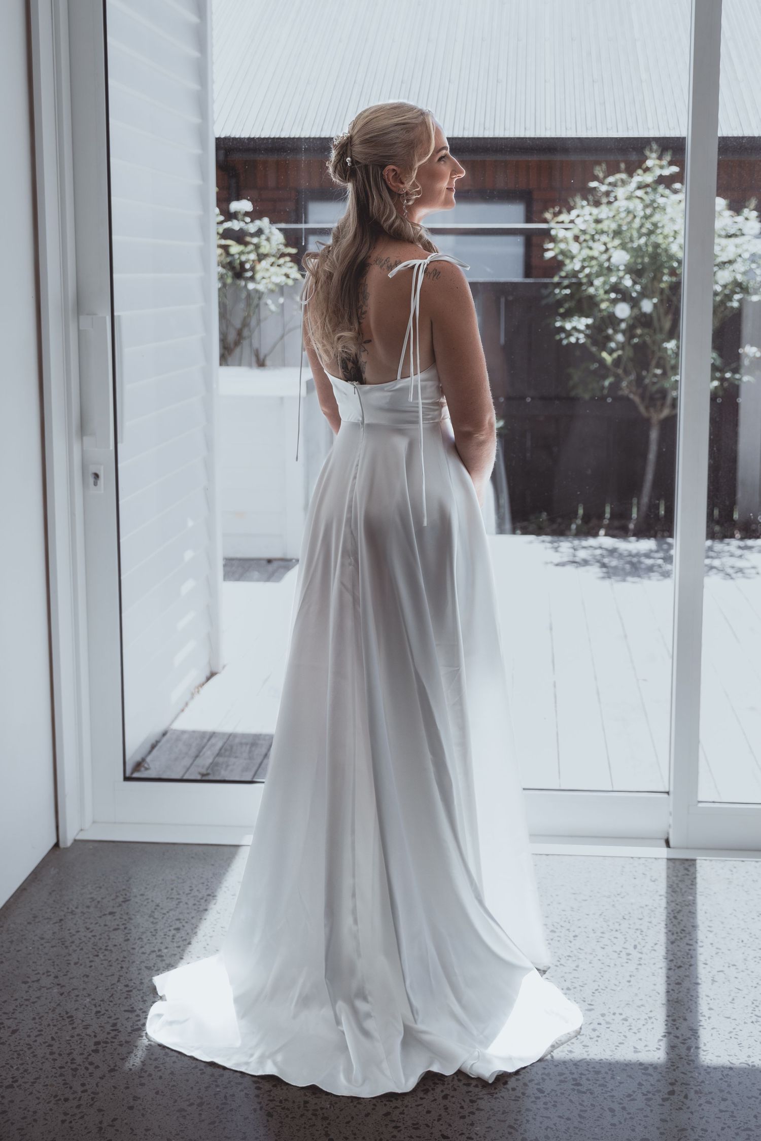 Bride in elegant white spaghetti strap wedding gown with flowing skirt stands by glass door looking outside.