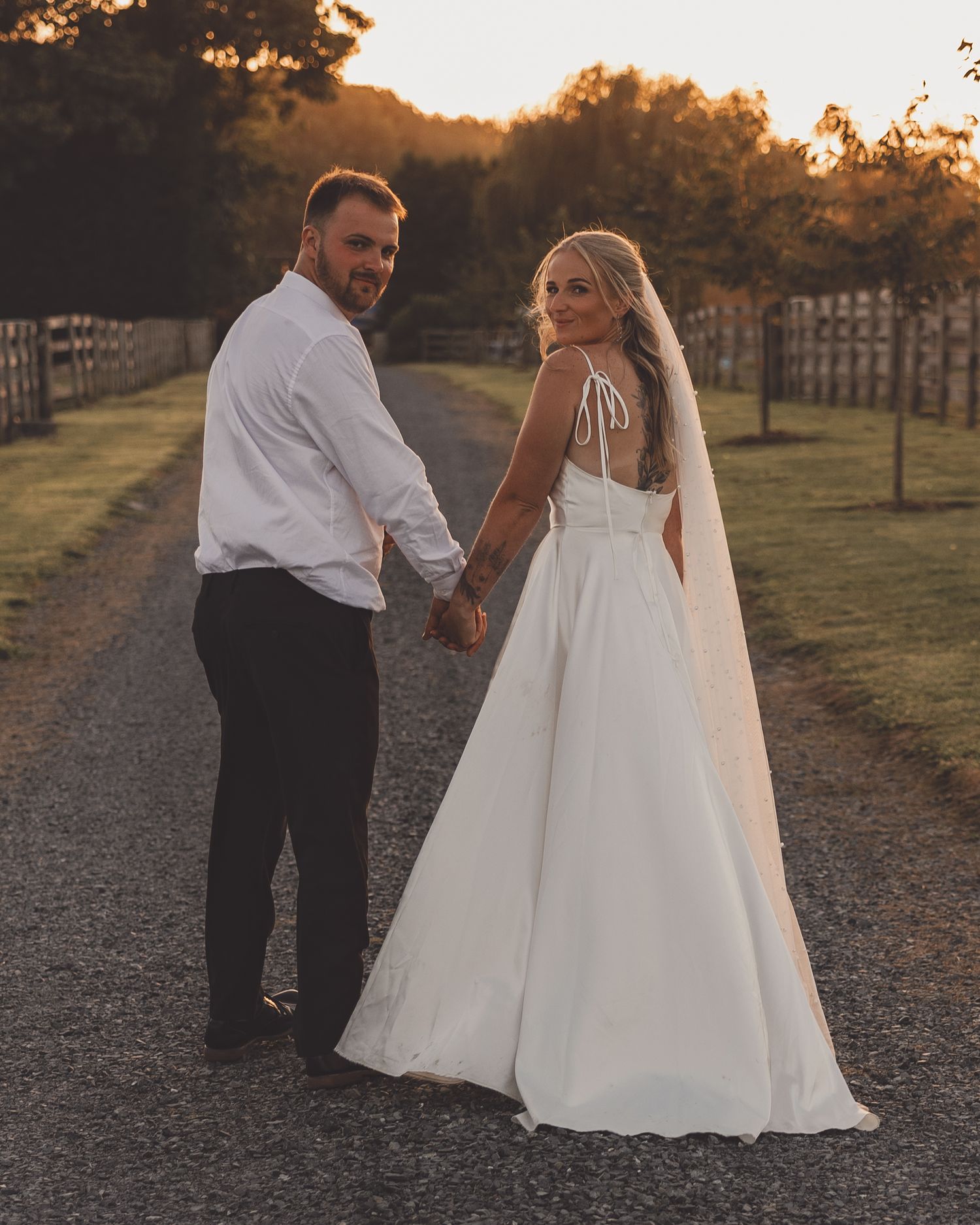 Bride and groom hold hands on a tree-lined path at golden hour, bride in elegant white gown with veil.