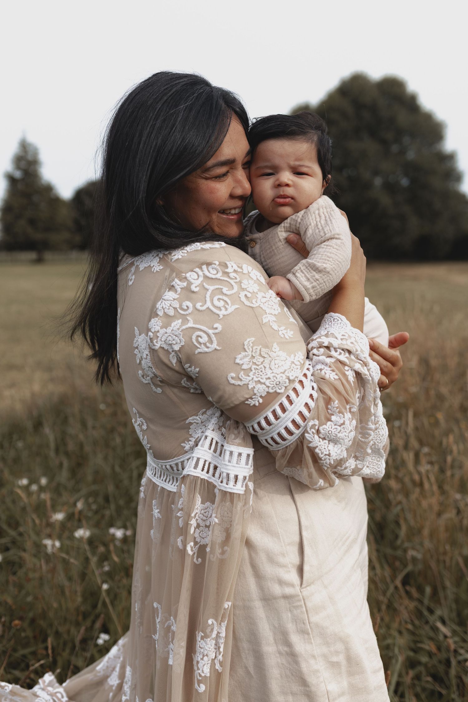 Mother in lace boho dress lovingly holding newborn baby in open field.