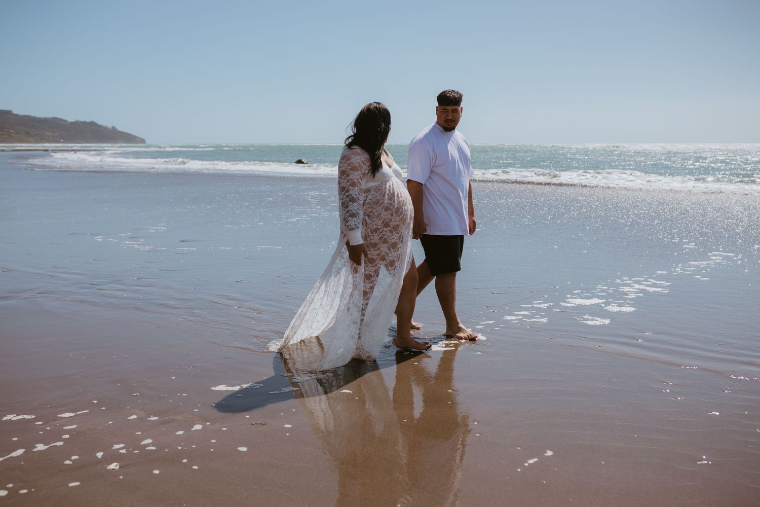 Couple walking along a sunny beach shoreline during a maternity photoshoot.