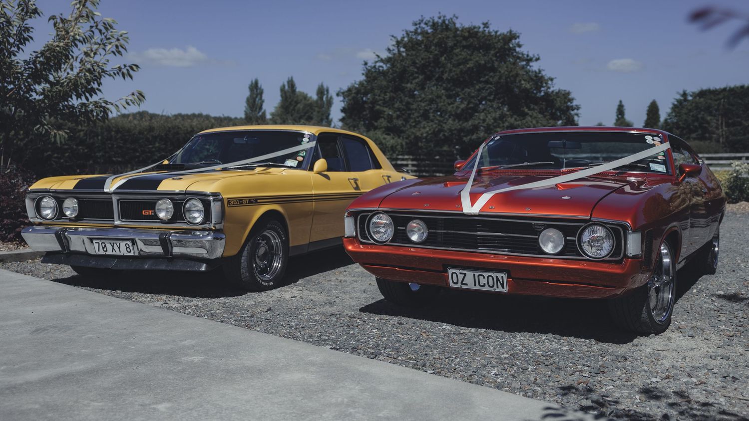 Two classic muscle cars, yellow and red, parked side by side decorated with white wedding ribbons.
