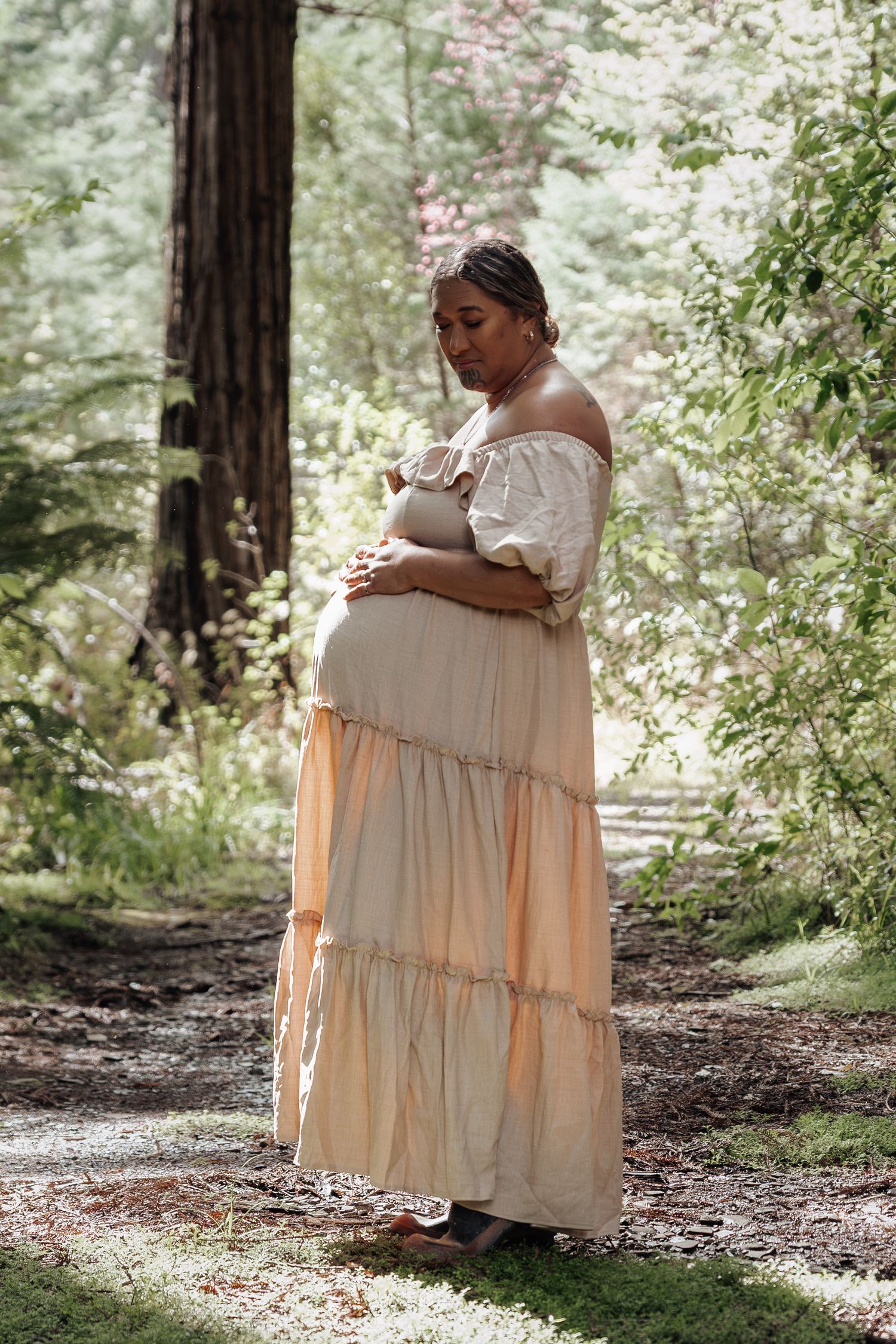 Pregnant woman in a flowing peach off-shoulder maxi dress standing in a sunlit forest, cradling her baby bump.
