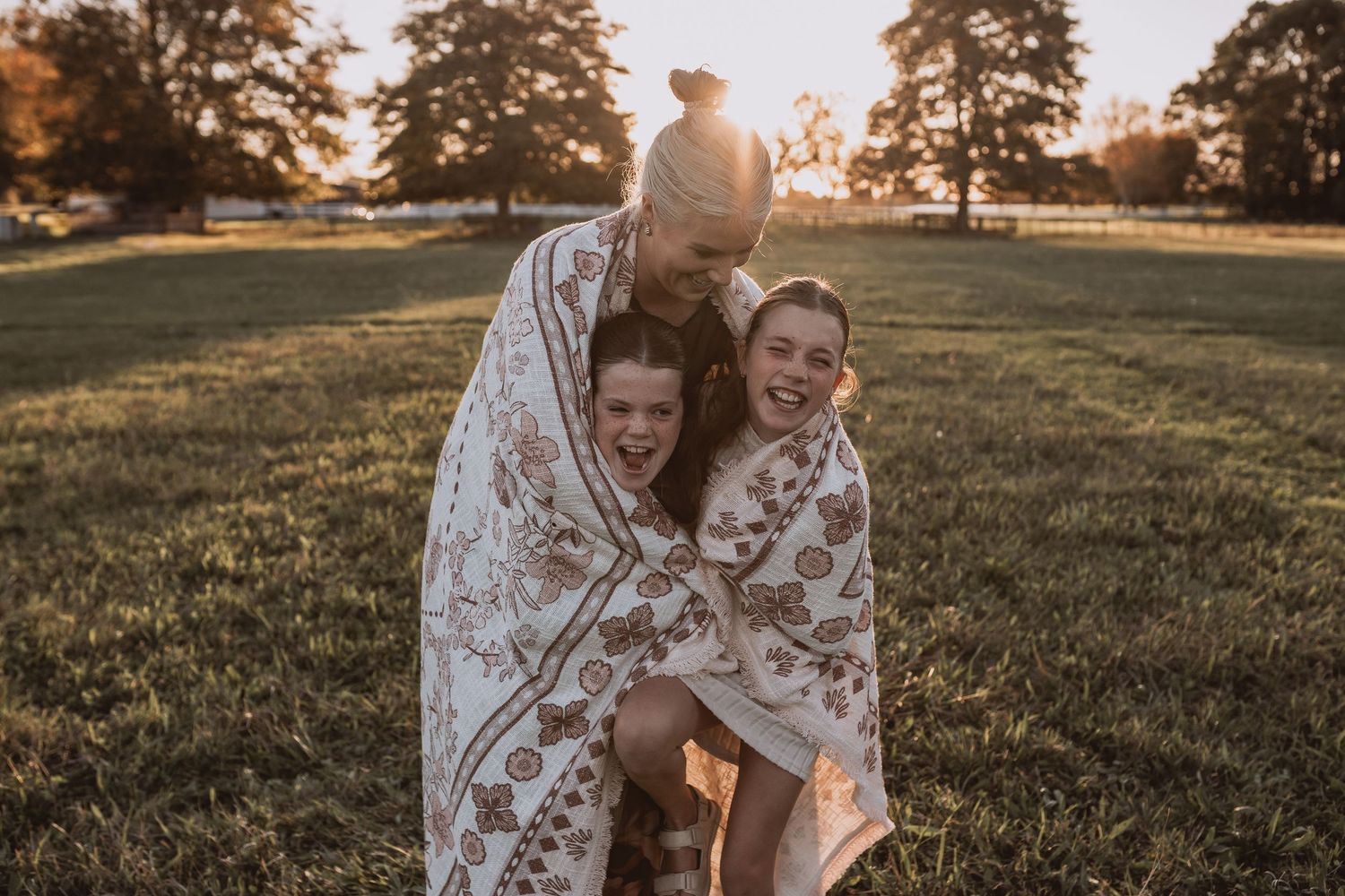 Joyful mother and two daughters wrapped together in boho floral blanket laughing at sunset outdoors.