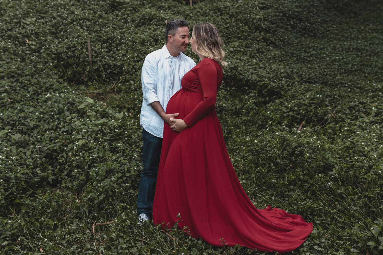 Couple sharing a tender moment outdoors, pregnant woman in elegant red gown embraced by partner.