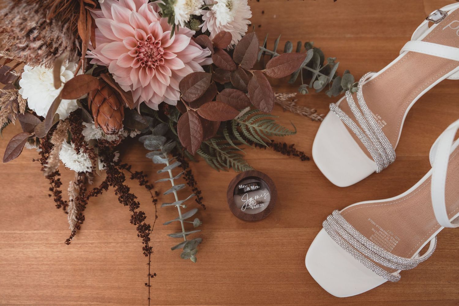 White bridal heels and wedding rings beside a rustic bouquet of pink dahlias and autumn foliage on wood.
