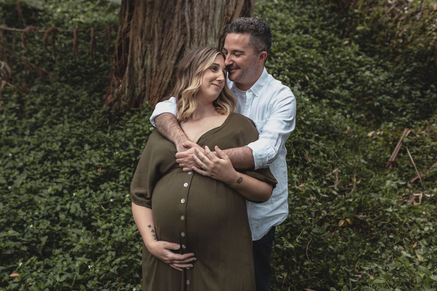 Pregnant woman in olive green dress embraced from behind by partner near a large tree.