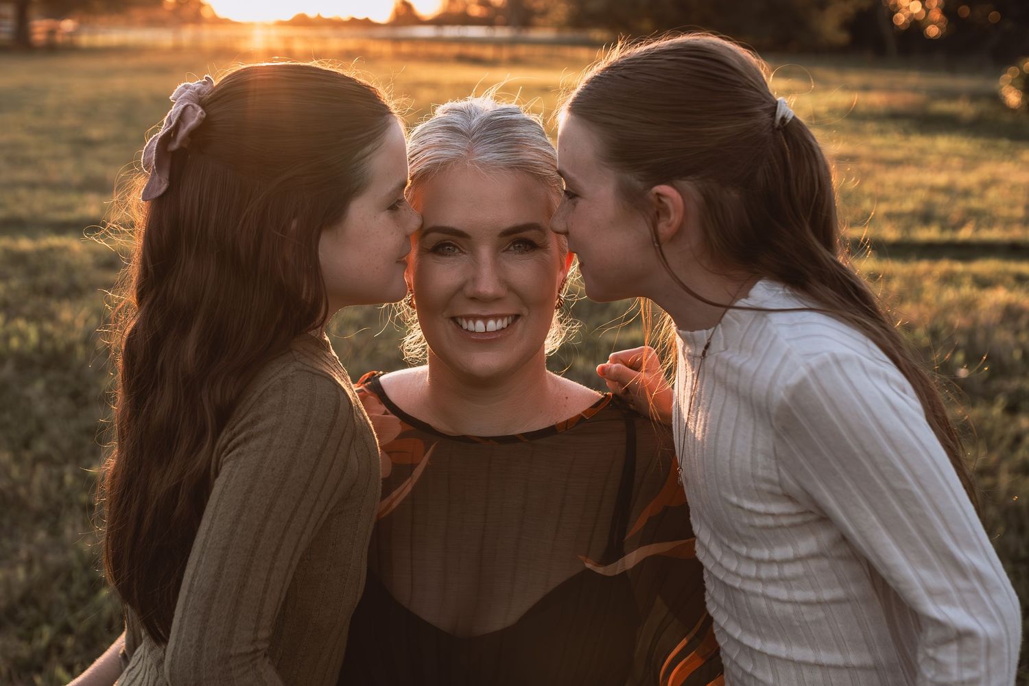 Two daughters kissing smiling mother on cheeks during warm sunset family photo session outdoors.