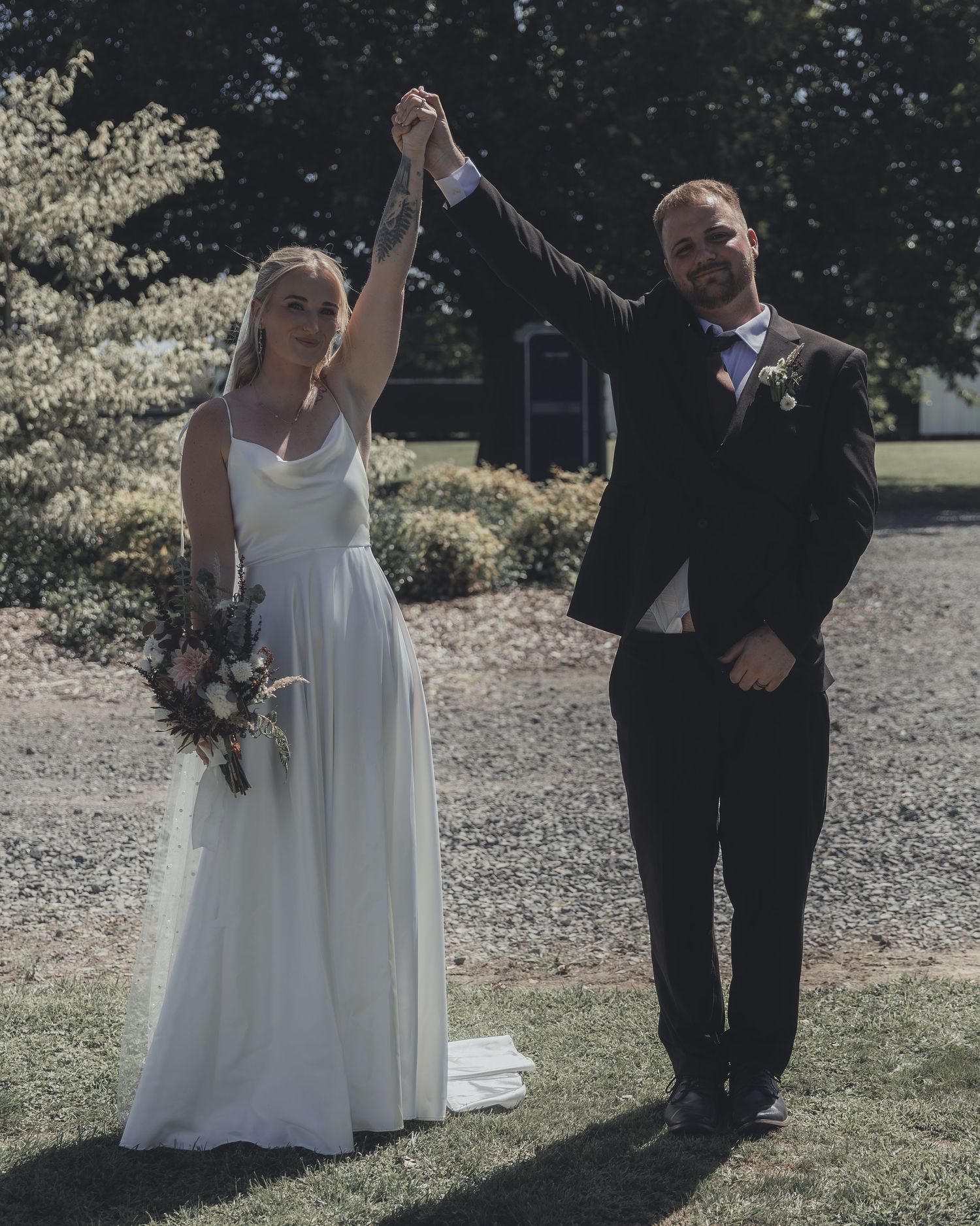 Bride and groom raising joined hands outdoors on their wedding day, bride in white gown holding bouquet, groom in black suit.