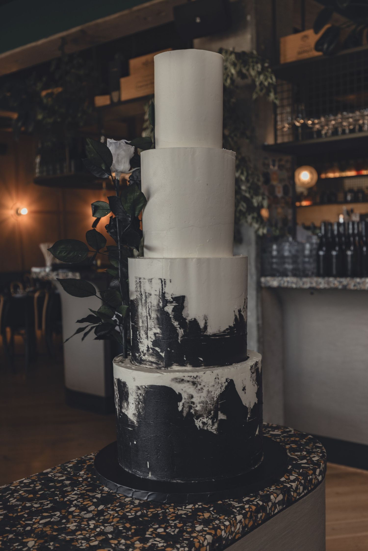 Four-tier black and white wedding cake with floral accents in moody bar setting.