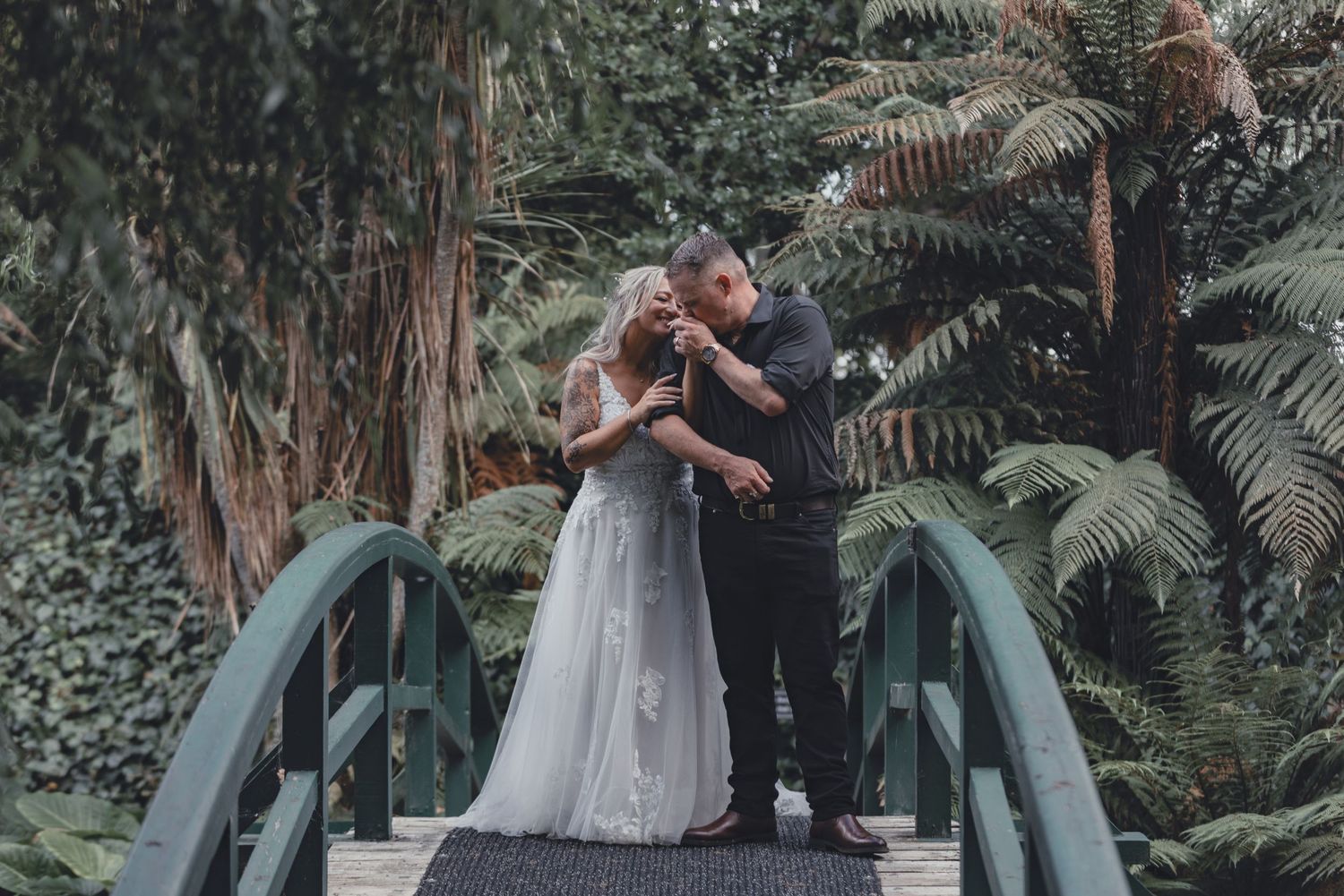 Bride and groom share romantic embrace on green bridge surrounded by lush ferns.