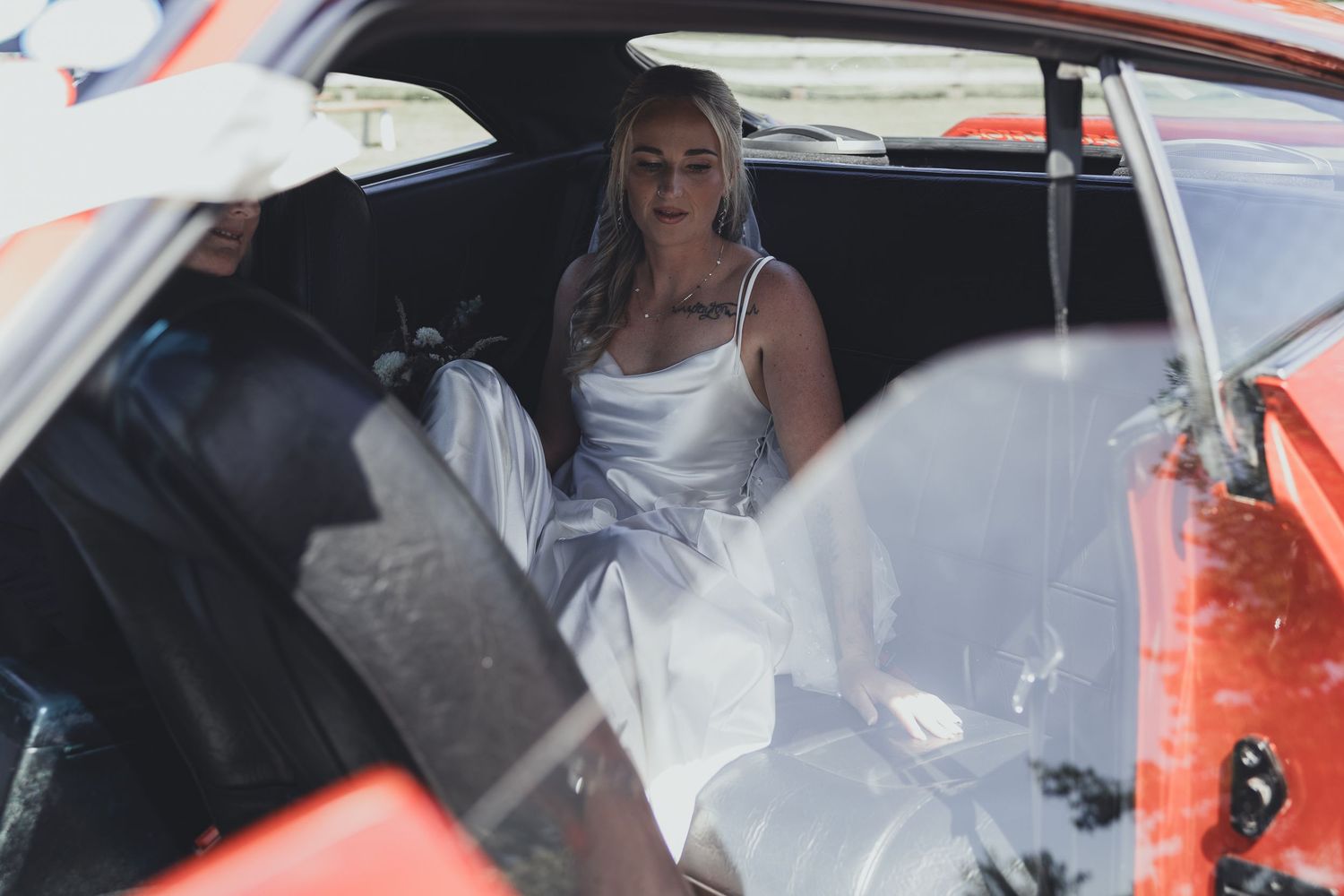 Blonde bride in a white satin wedding dress sitting inside a vintage red car on her wedding day.