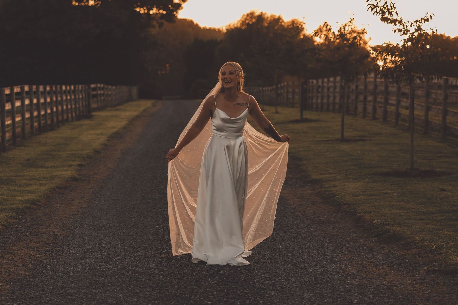 Bride in elegant white gown holding dress hem on gravel path at golden sunset, flanked by wooden fences.