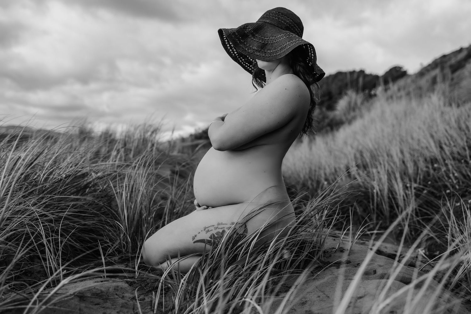 Elegant black and white maternity photo of pregnant woman in wide-brim hat among tall grass.