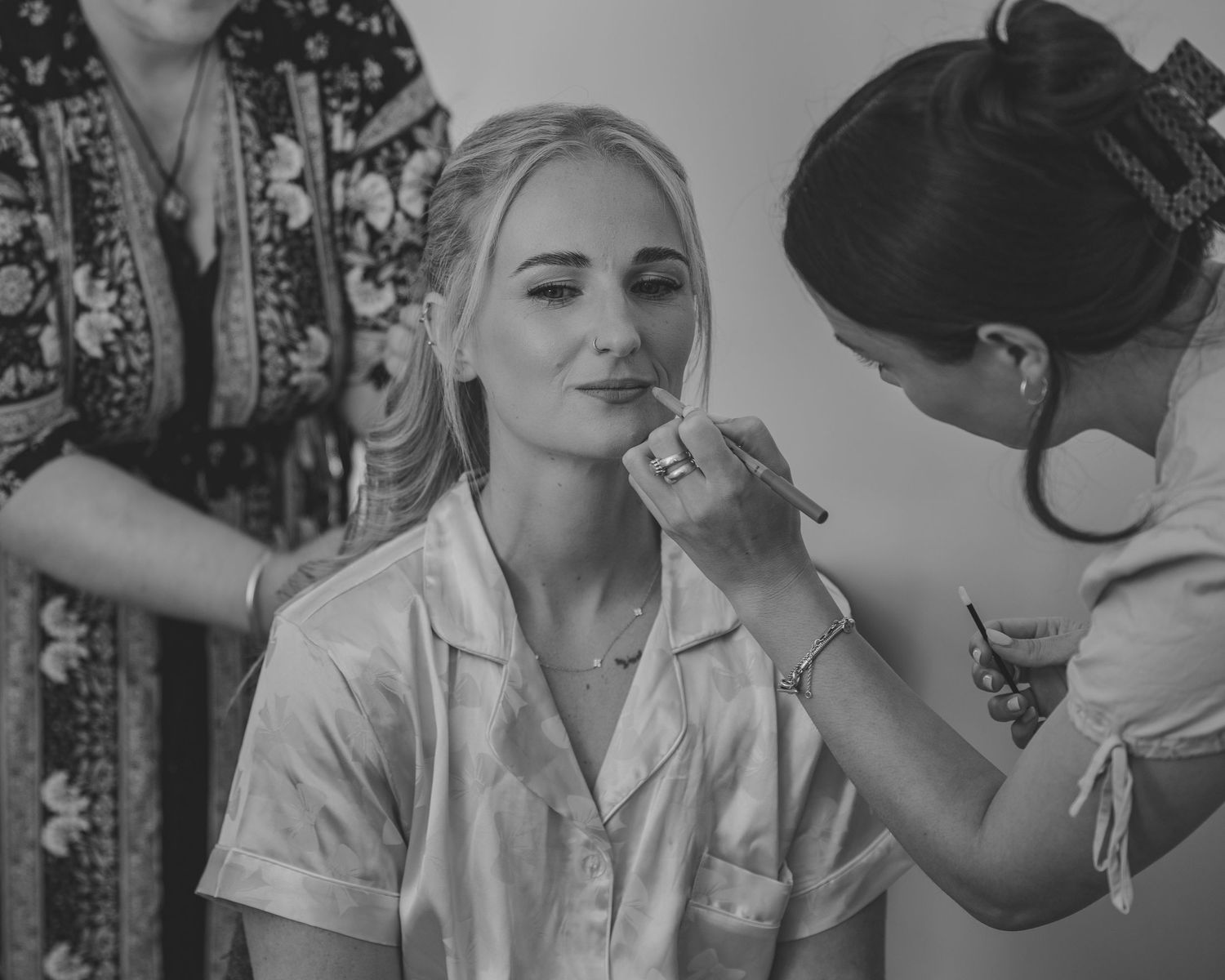 Bride getting makeup applied by artist while hair is styled in black and white wedding preparation photo.