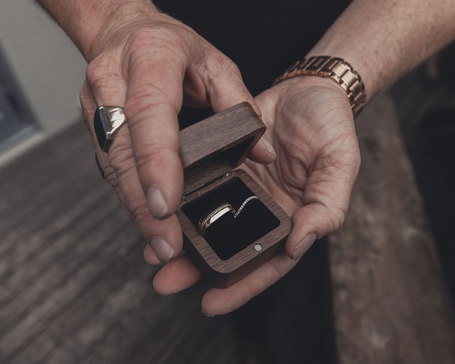 Hands holding wooden ring box with gold wedding band and bracelet visible.