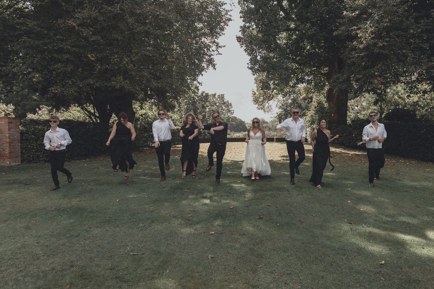 Wedding party jumps joyfully together on green lawn surrounded by trees.