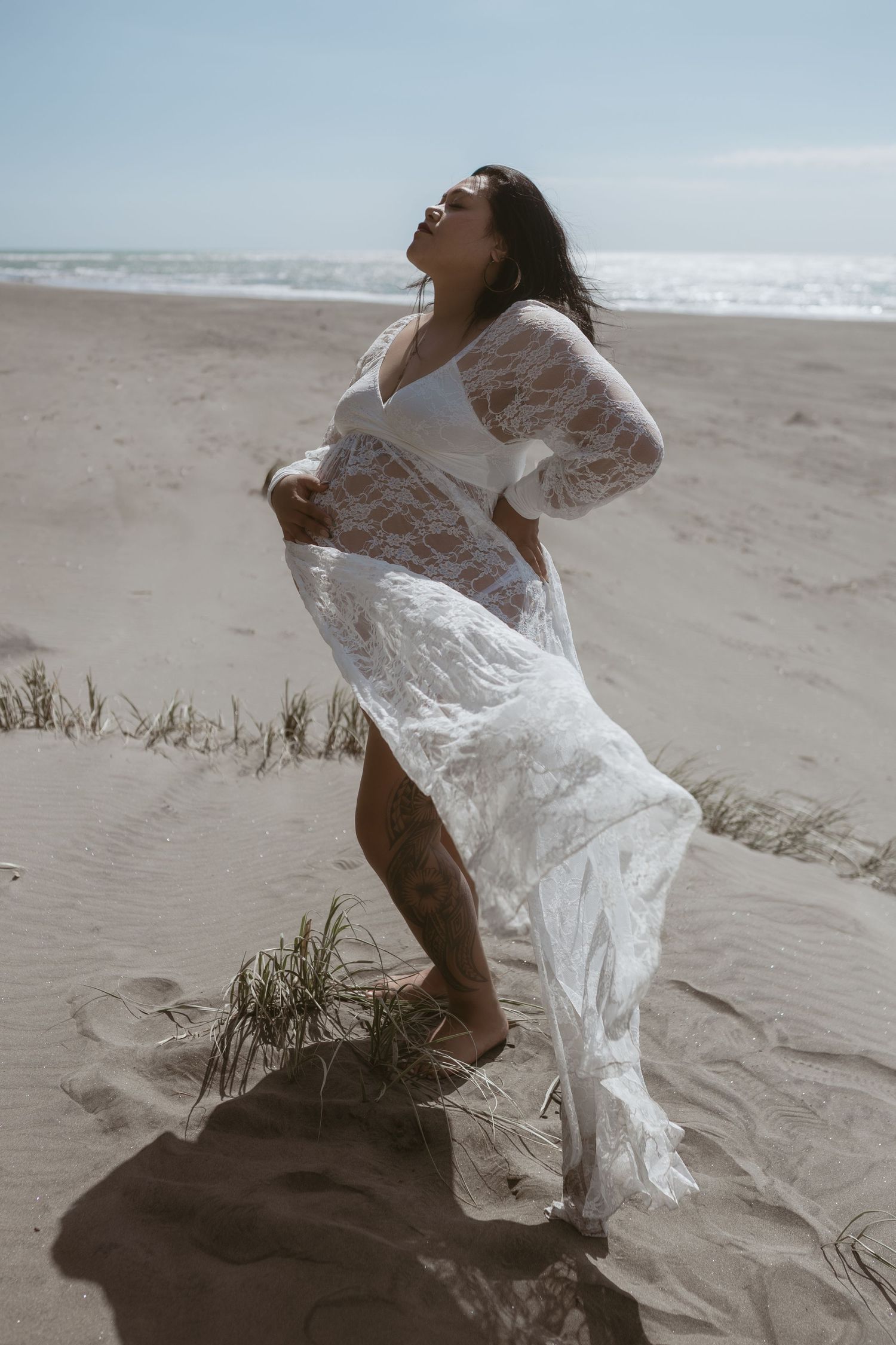 Pregnant woman in flowing white lace dress posing on sandy beach dunes with ocean in background.