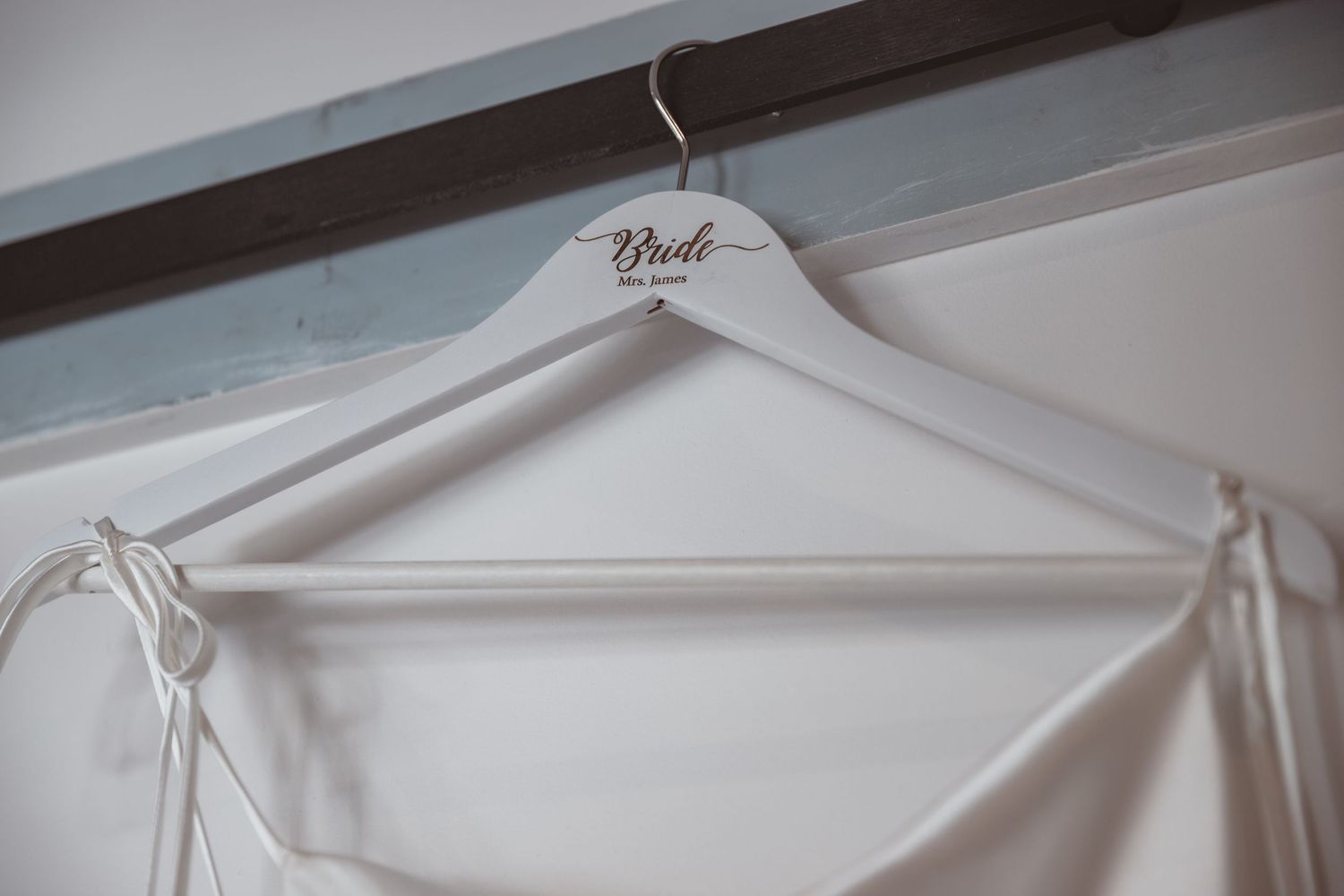 White wooden bride hanger with cursive script displayed on rustic wooden rod with wedding dress.