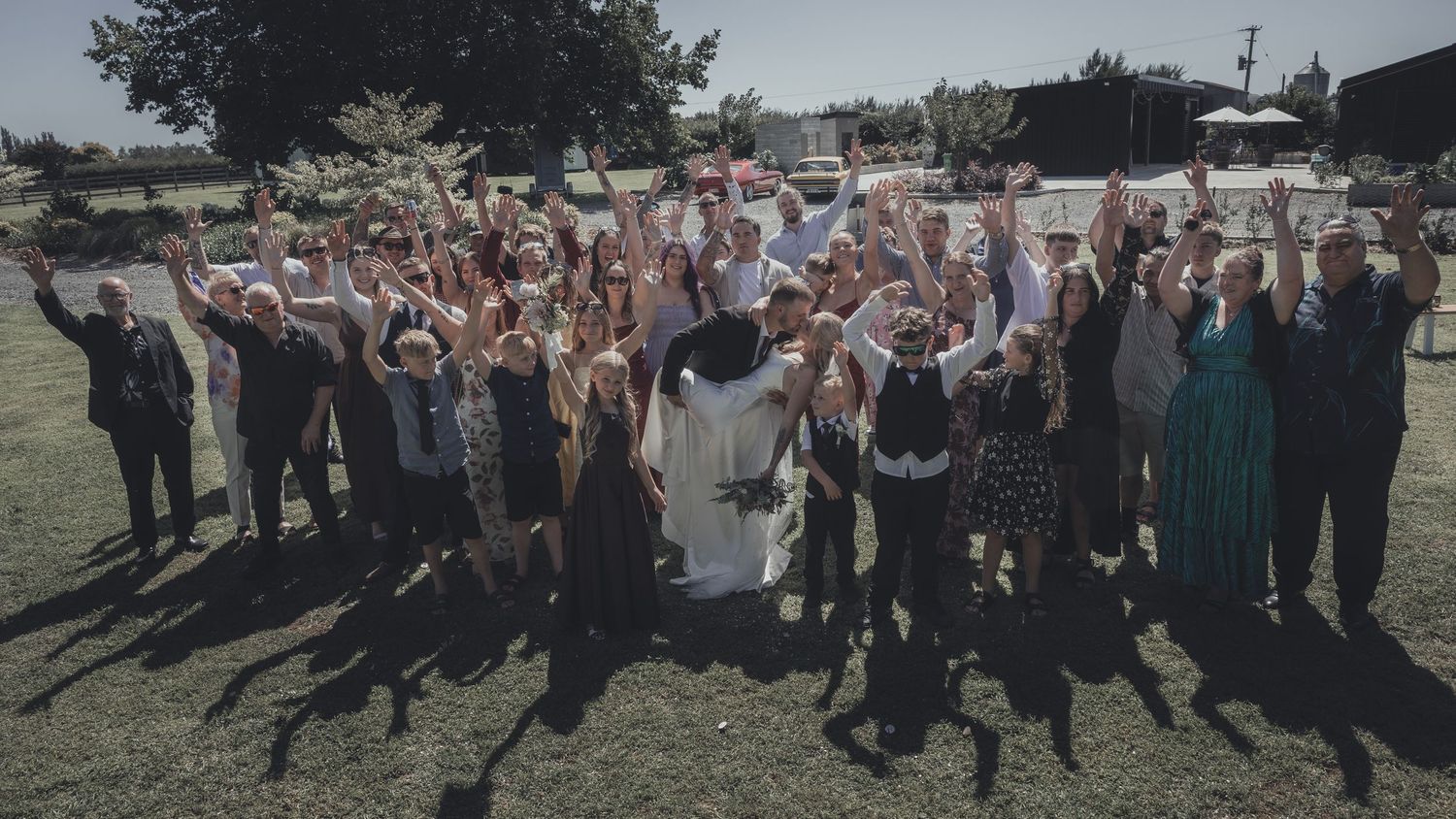 Wedding group photo with bride and groom surrounded by cheering guests with raised arms on a sunny day.