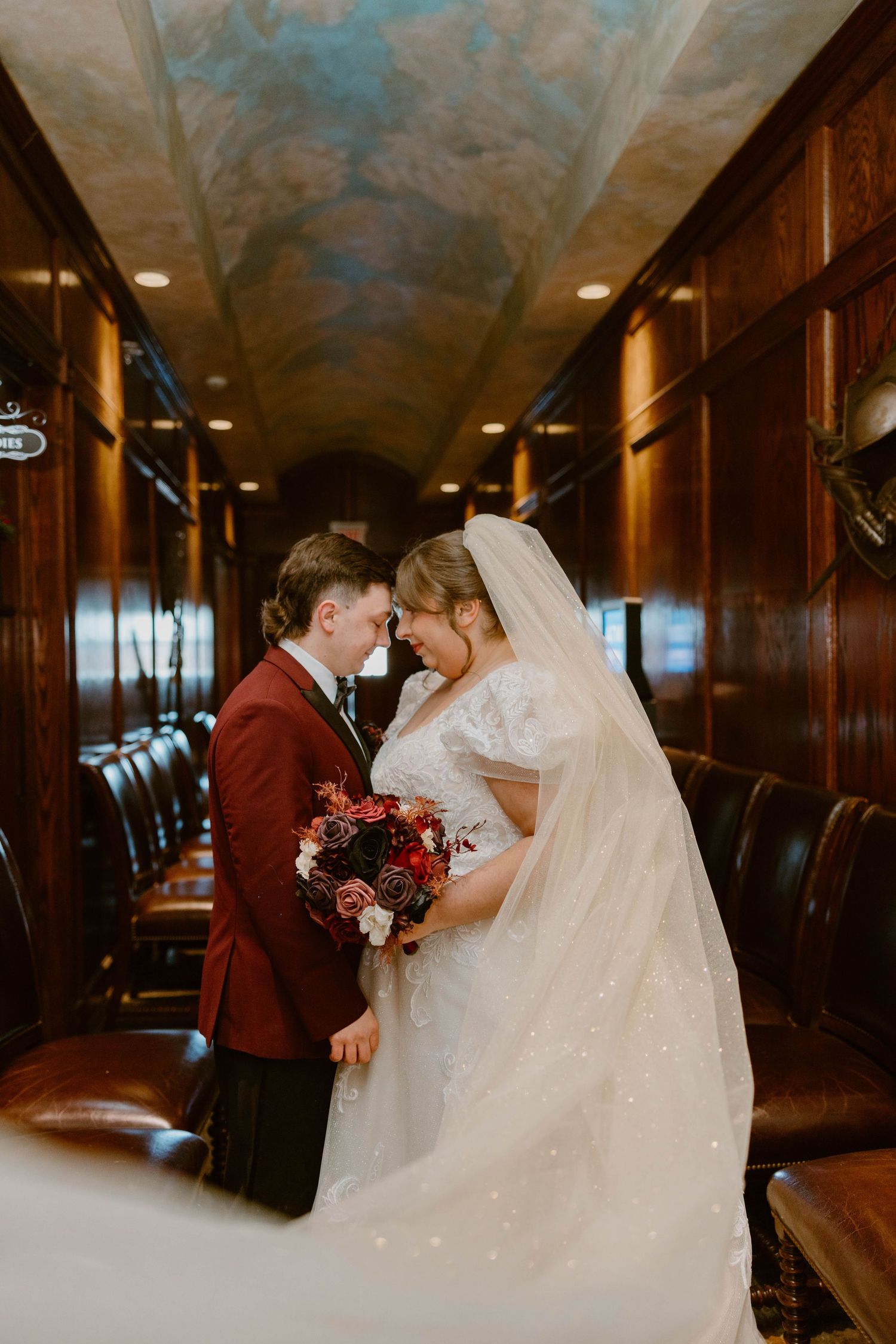 Bride and groom touching foreheads in elegant wood-paneled room with painted ceiling and red rose bouquet at Shakespeare's Restaurant