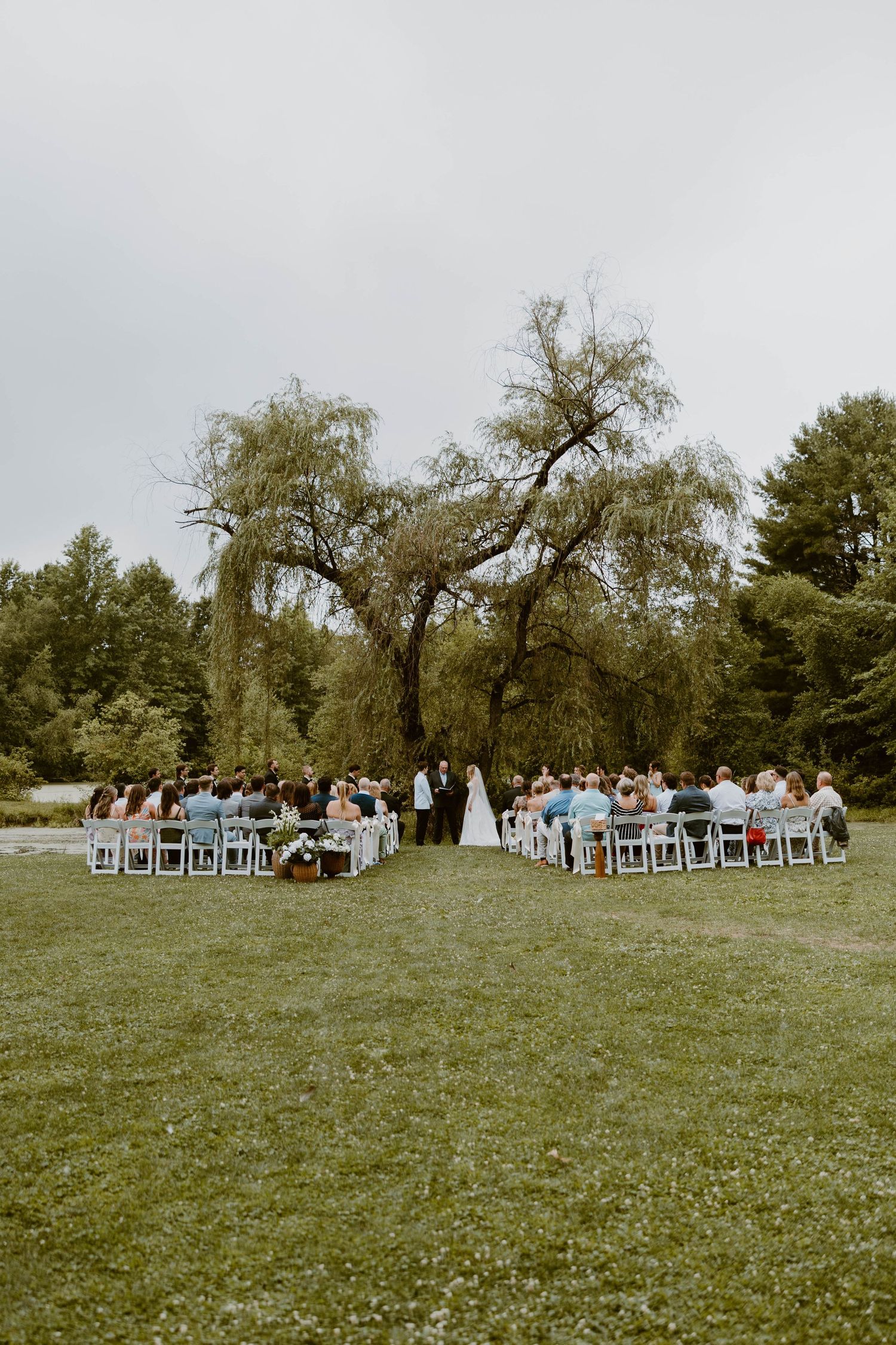 Outdoor wedding ceremony with guests seated in white chairs beneath a large weeping willow tree on green lawn.
