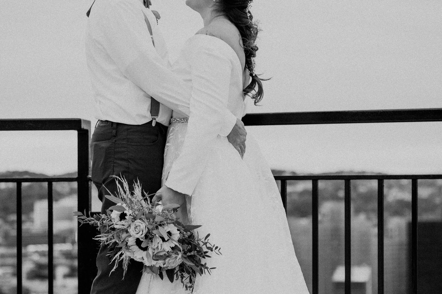 Black and white photo of bride and groom embracing on a rooftop balcony with city views below.