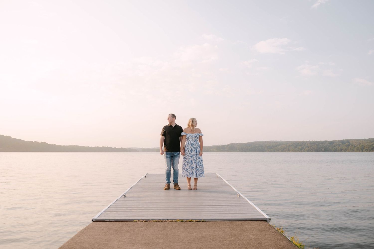 Couple standing side by side on a wooden dock extending into a calm lake at sunset, with hazy hills in the distance.