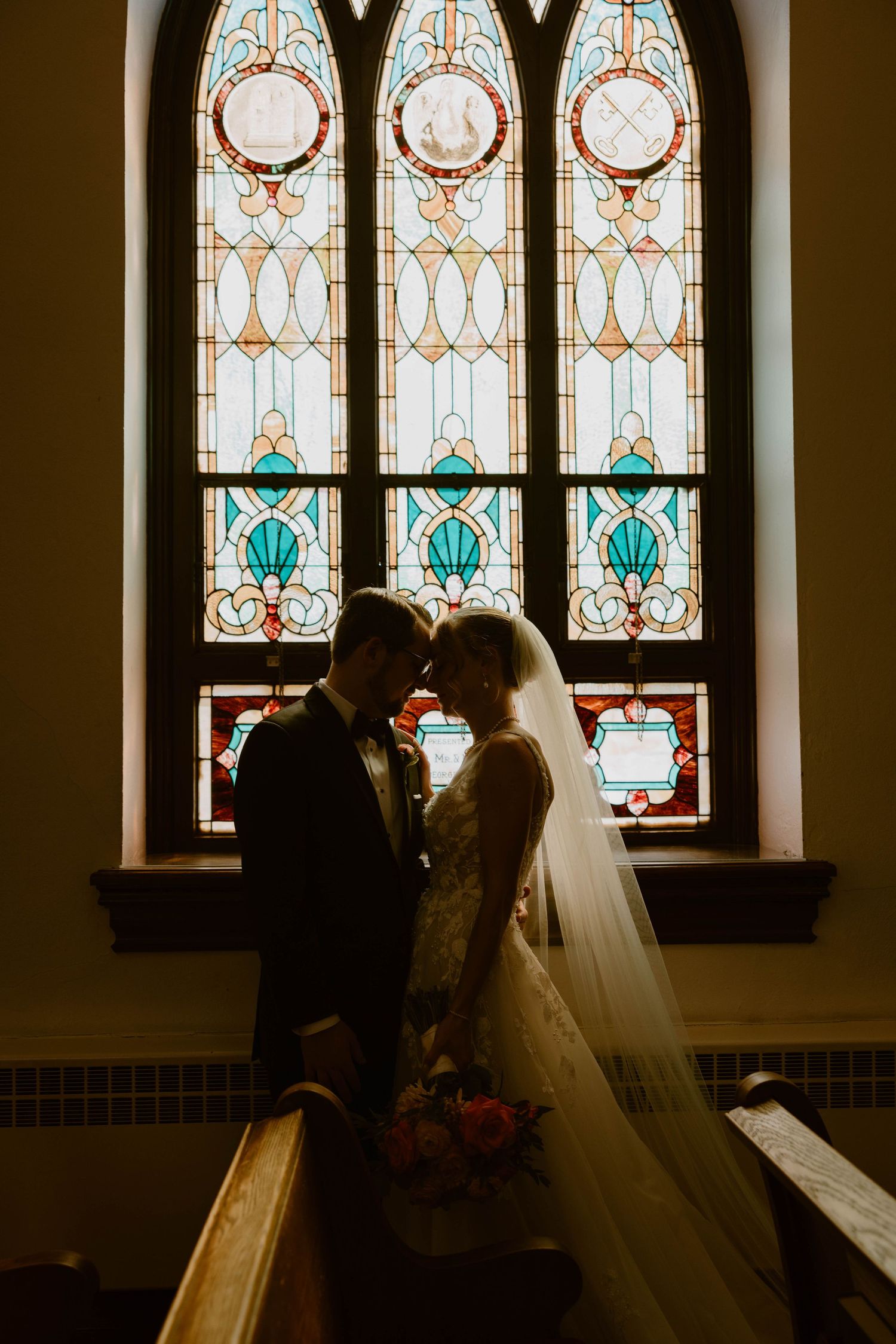 Bride and groom silhouetted against a stunning stained glass church window.
