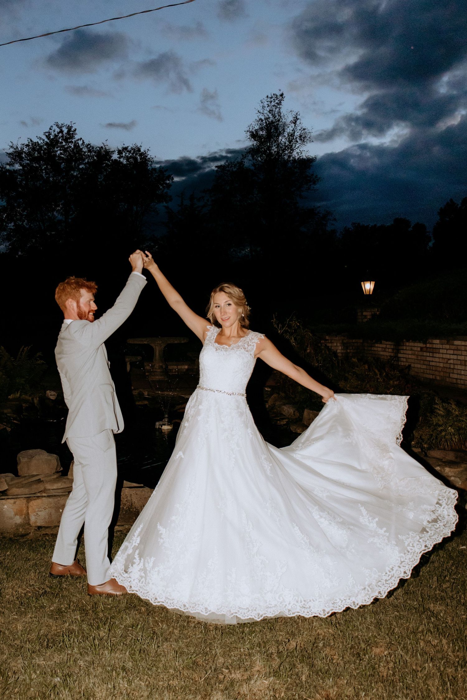 Bride and groom share a romantic dance outdoors at twilight, her white lace gown flowing gracefully.