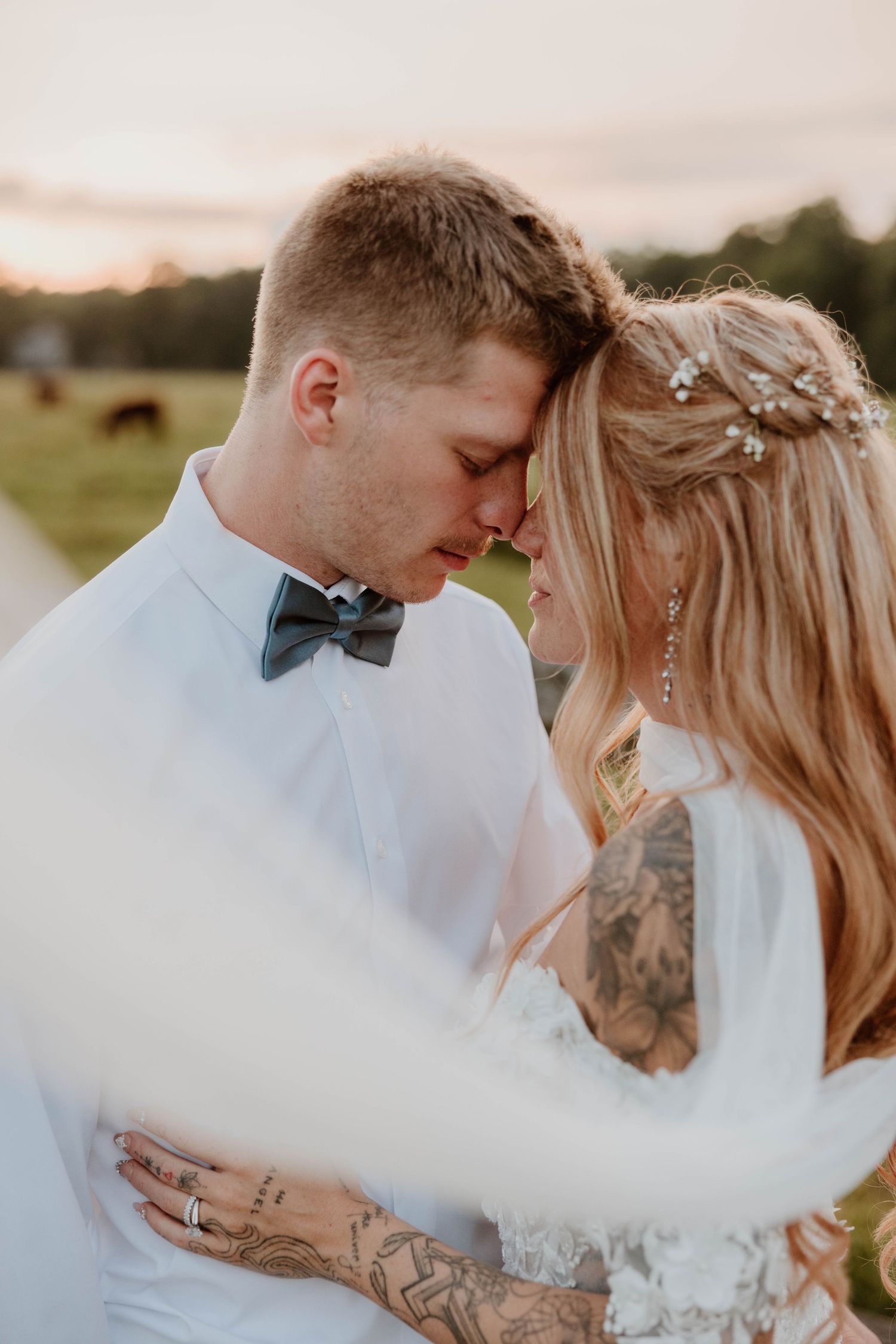 Tattooed bride and groom in white shirt with gray bow tie touching foreheads at golden sunset at Armstrong Farms in Butler County