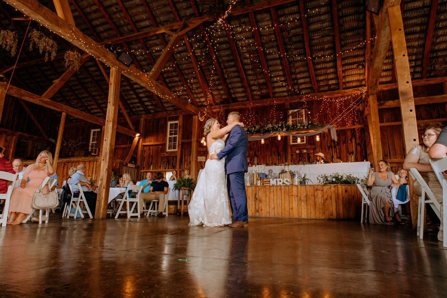 Bride and groom share their first dance inside a warmly lit rustic wooden barn wedding reception.