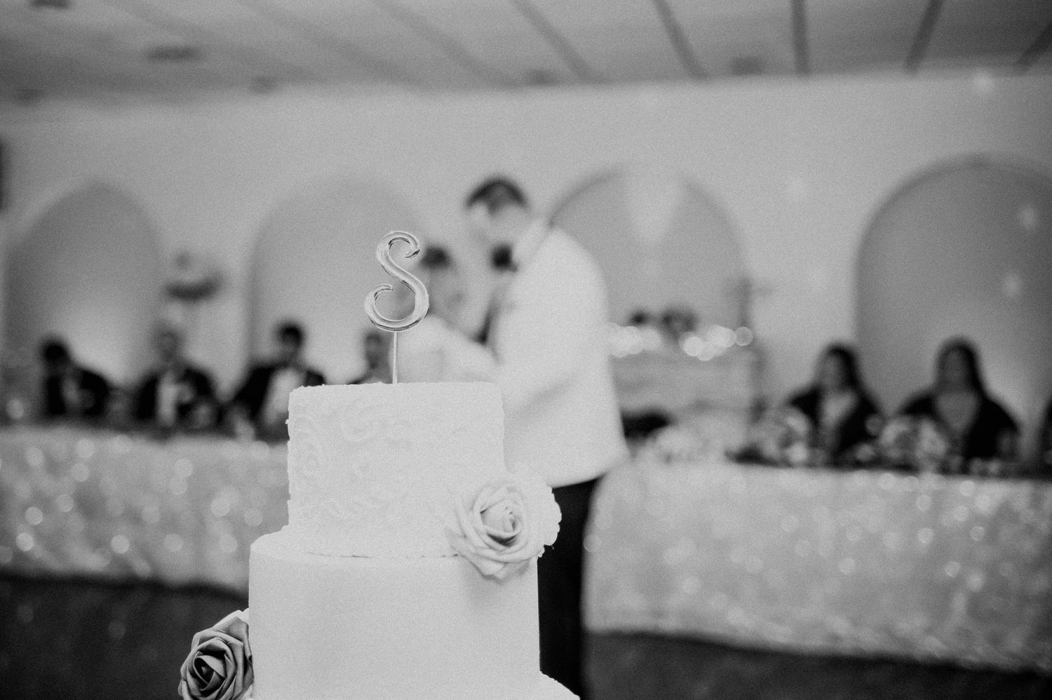 Black and white photo of a tiered white wedding cake with monogram topper at a reception.