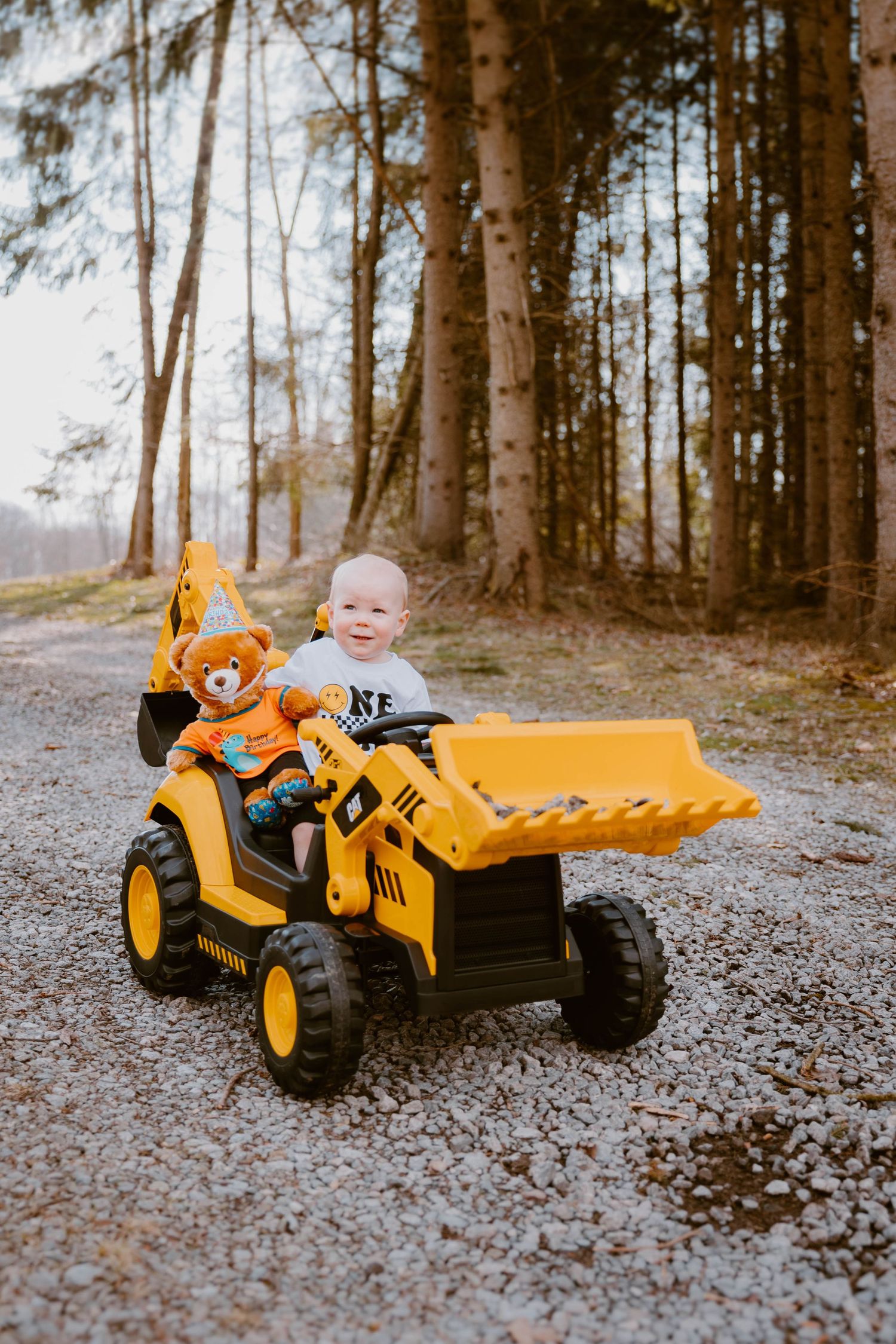 Happy toddler sits on a yellow ride-on toy excavator with a teddy bear on a gravel forest path.