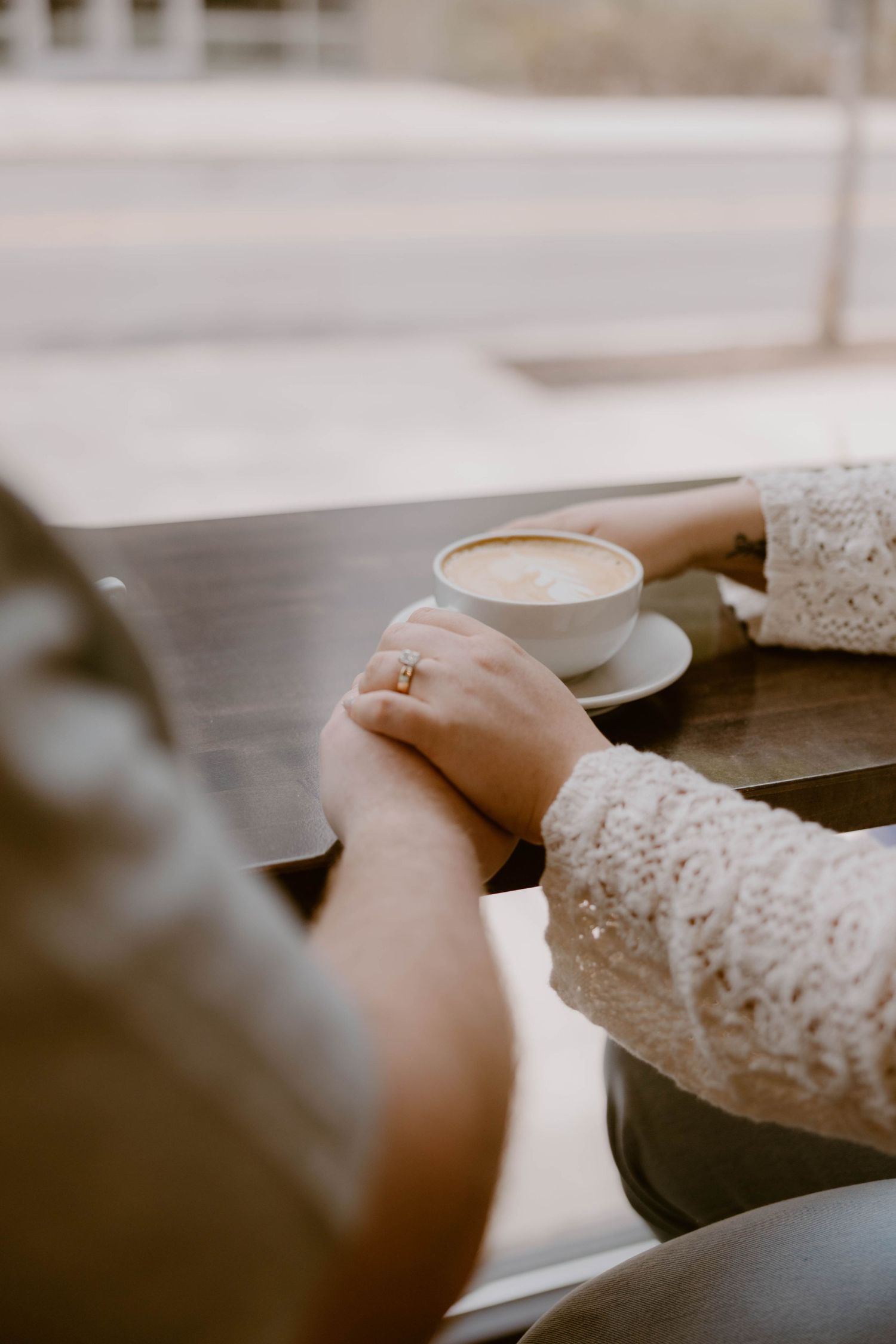 Couple holds hands at a cafe table beside a latte, with an engagement ring visible on her finger.