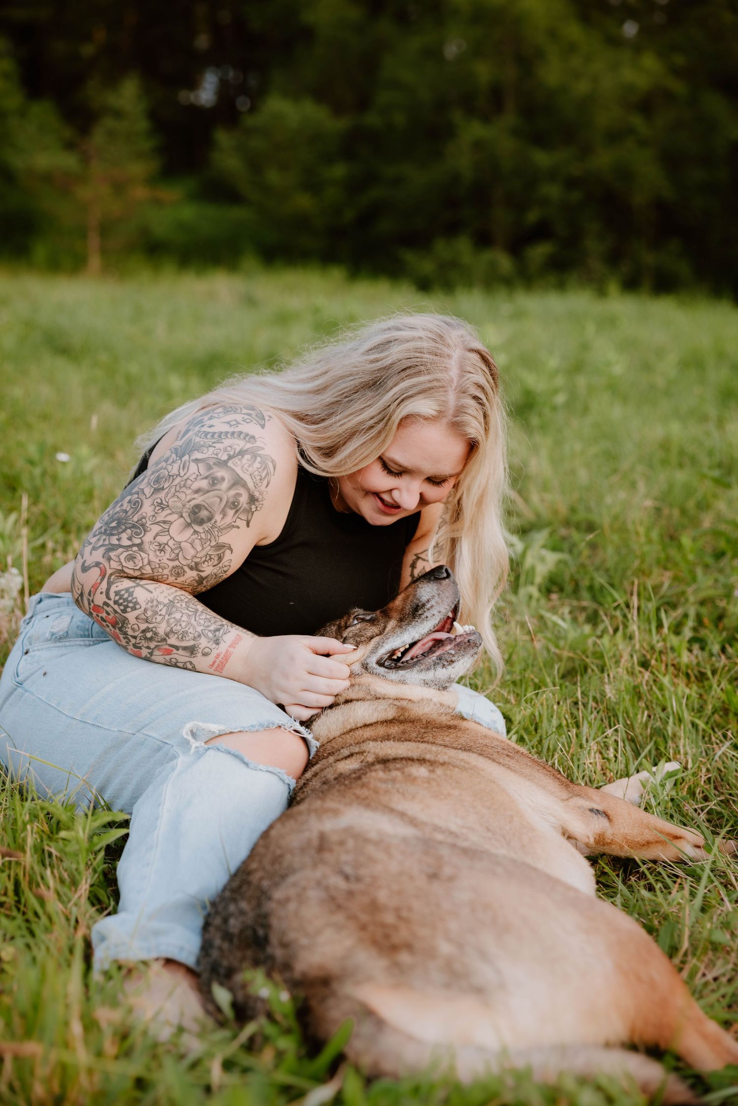 Tattooed blonde woman sitting in grass cuddling with her large dog in a sunny green field.