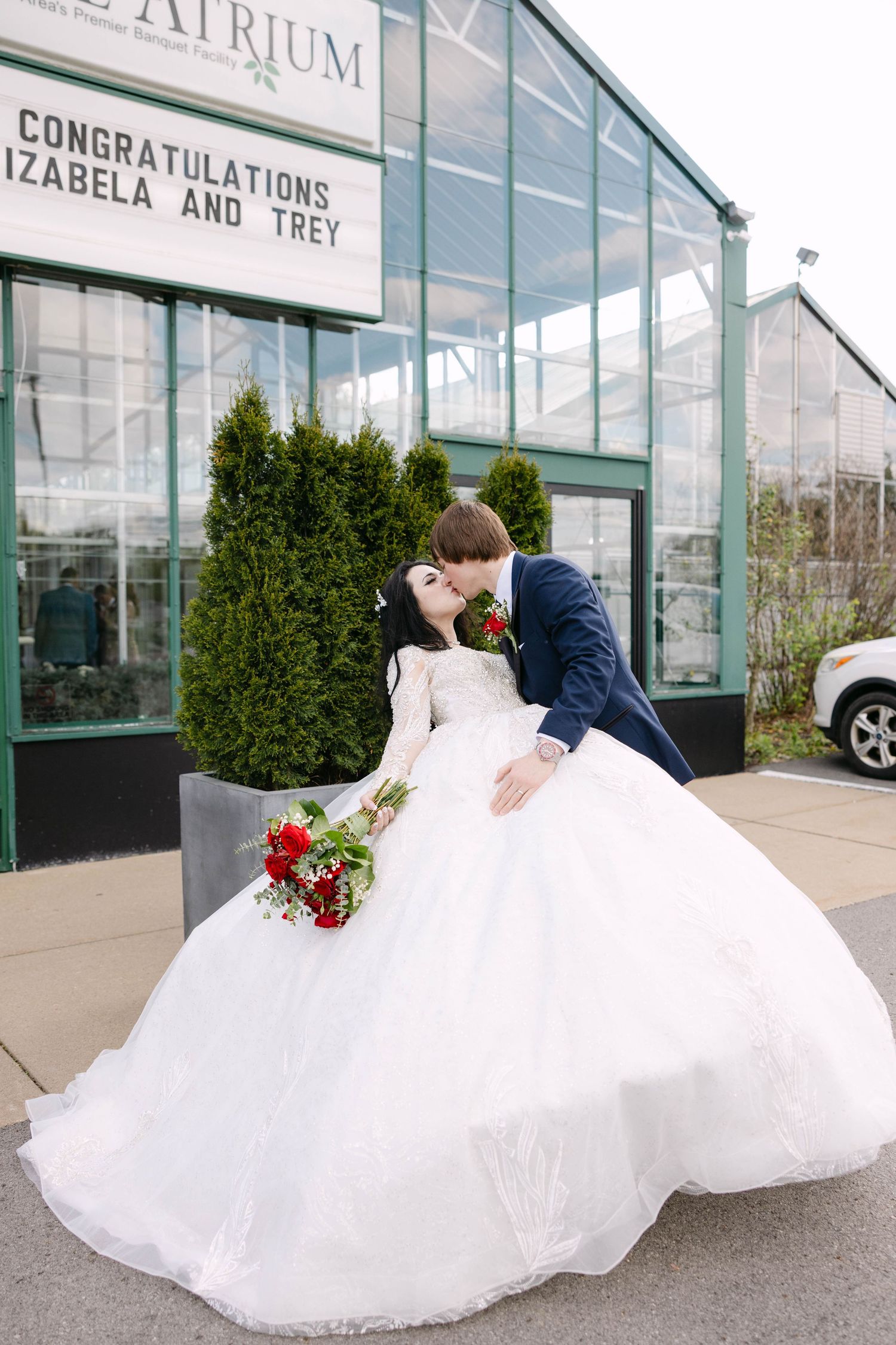Groom in navy suit dips and kisses bride in voluminous white lace gown outside The Atrium wedding venue.