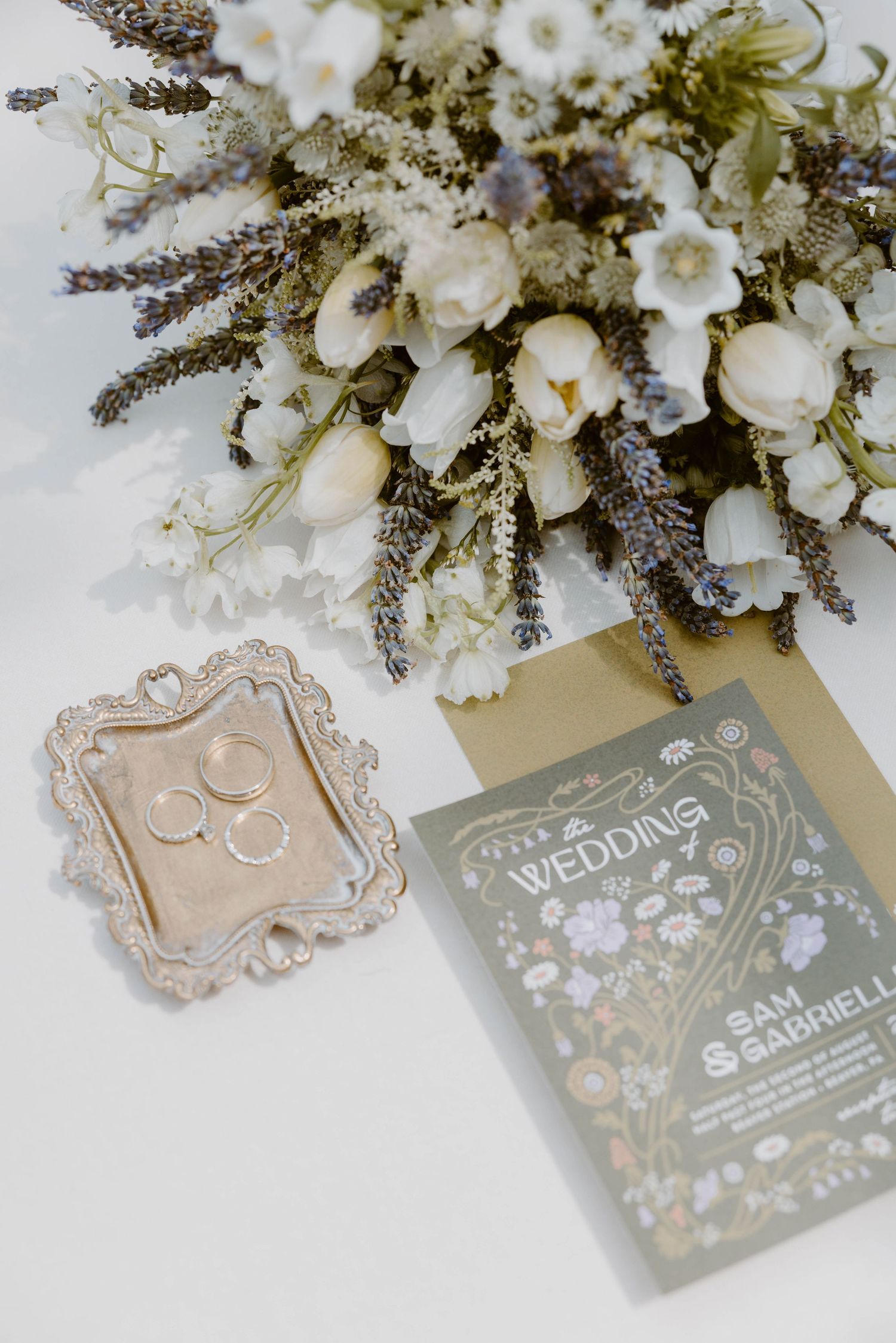 Wedding flat lay featuring white and lavender bouquet, gold rings on ornate tray, and floral invitation.
