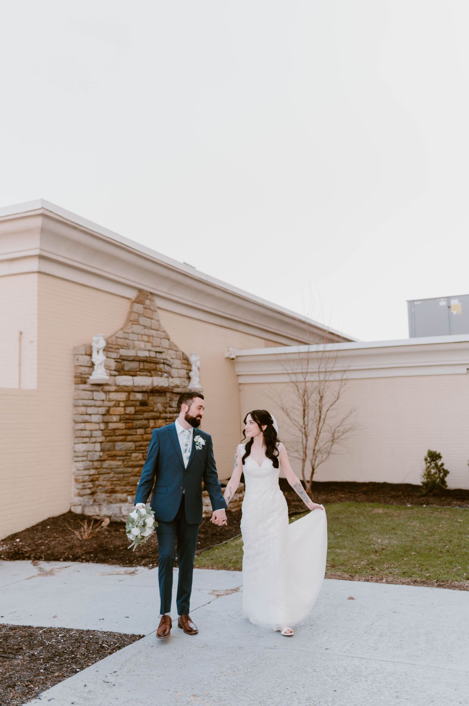 Bride and groom holding hands walking together outside a venue with a stone fireplace wall.