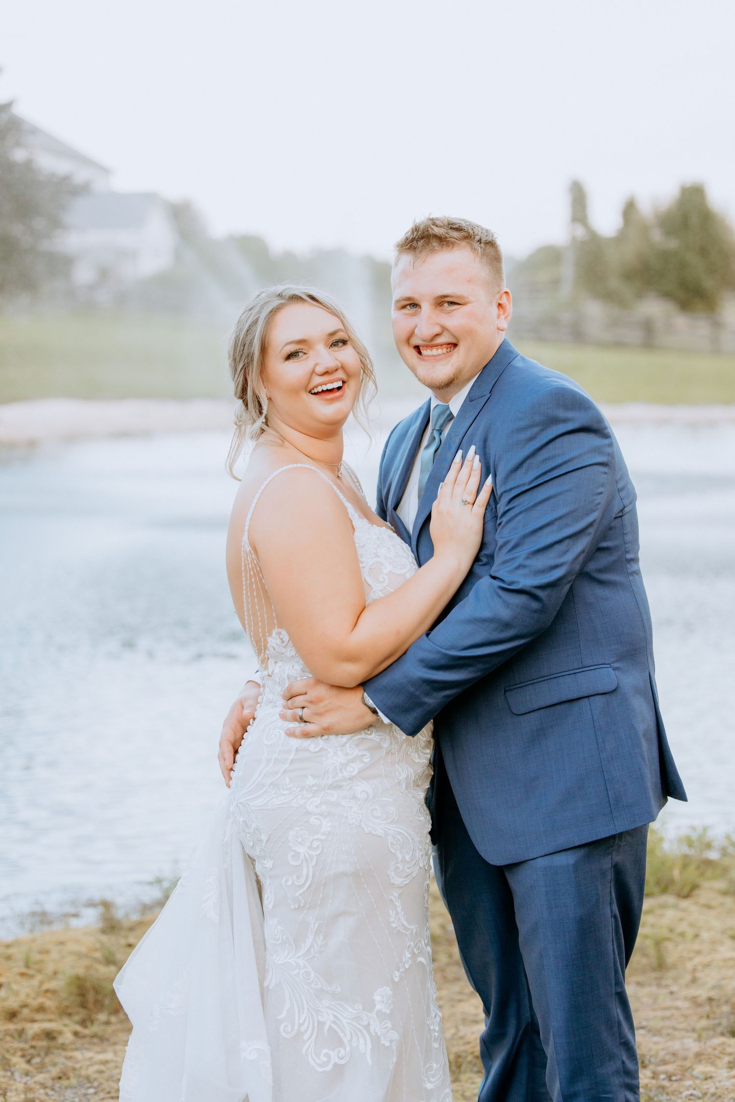 Joyful bride and groom embrace and smile together near a serene pond on their wedding day.