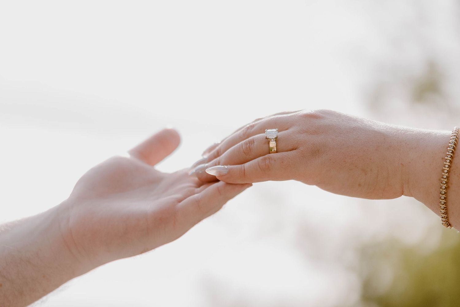 Close-up of two hands touching as one displays a delicate gold engagement ring against a soft blurred background.