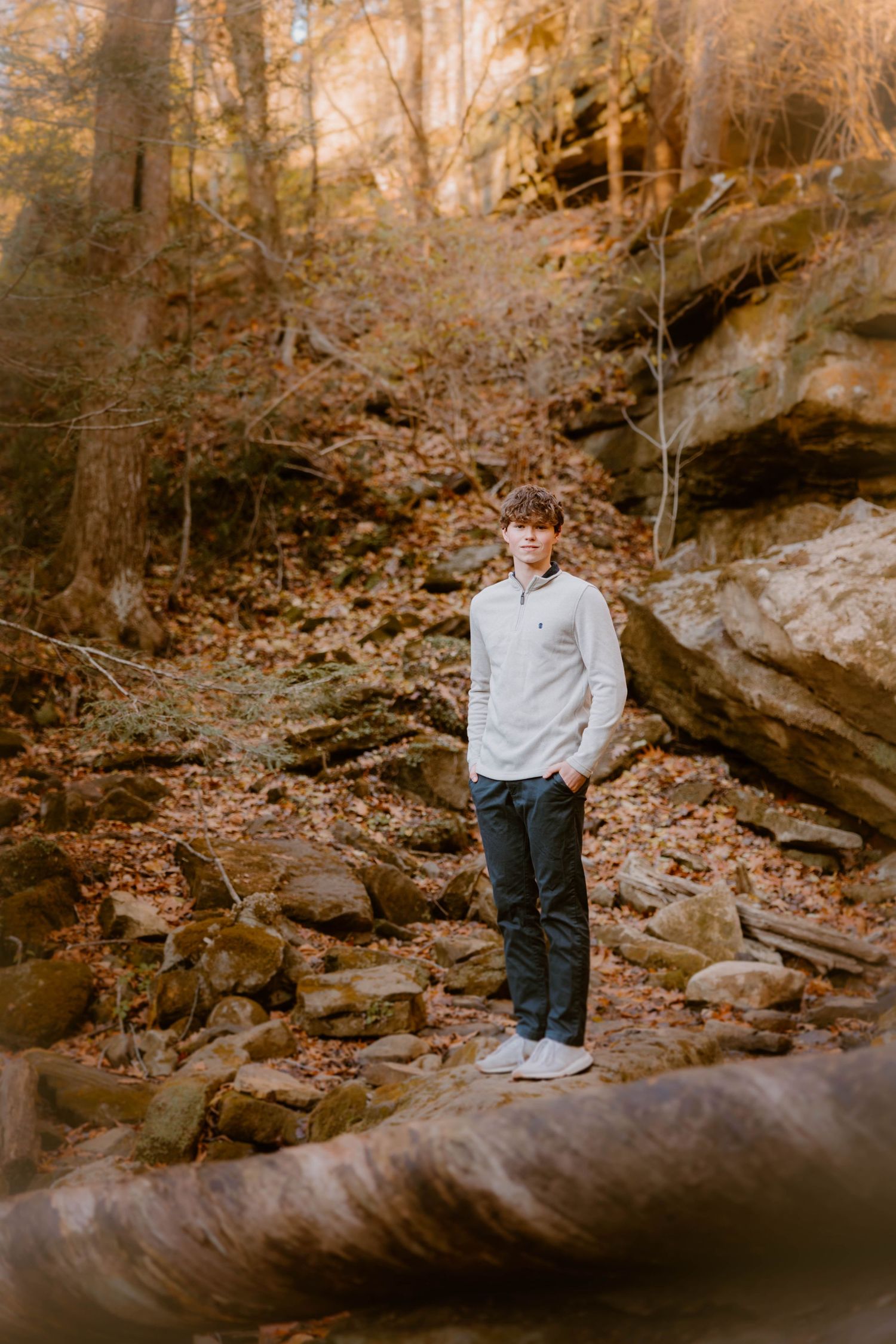 Teen boy in white sweater standing among autumn rocks and fallen leaves in a forest.