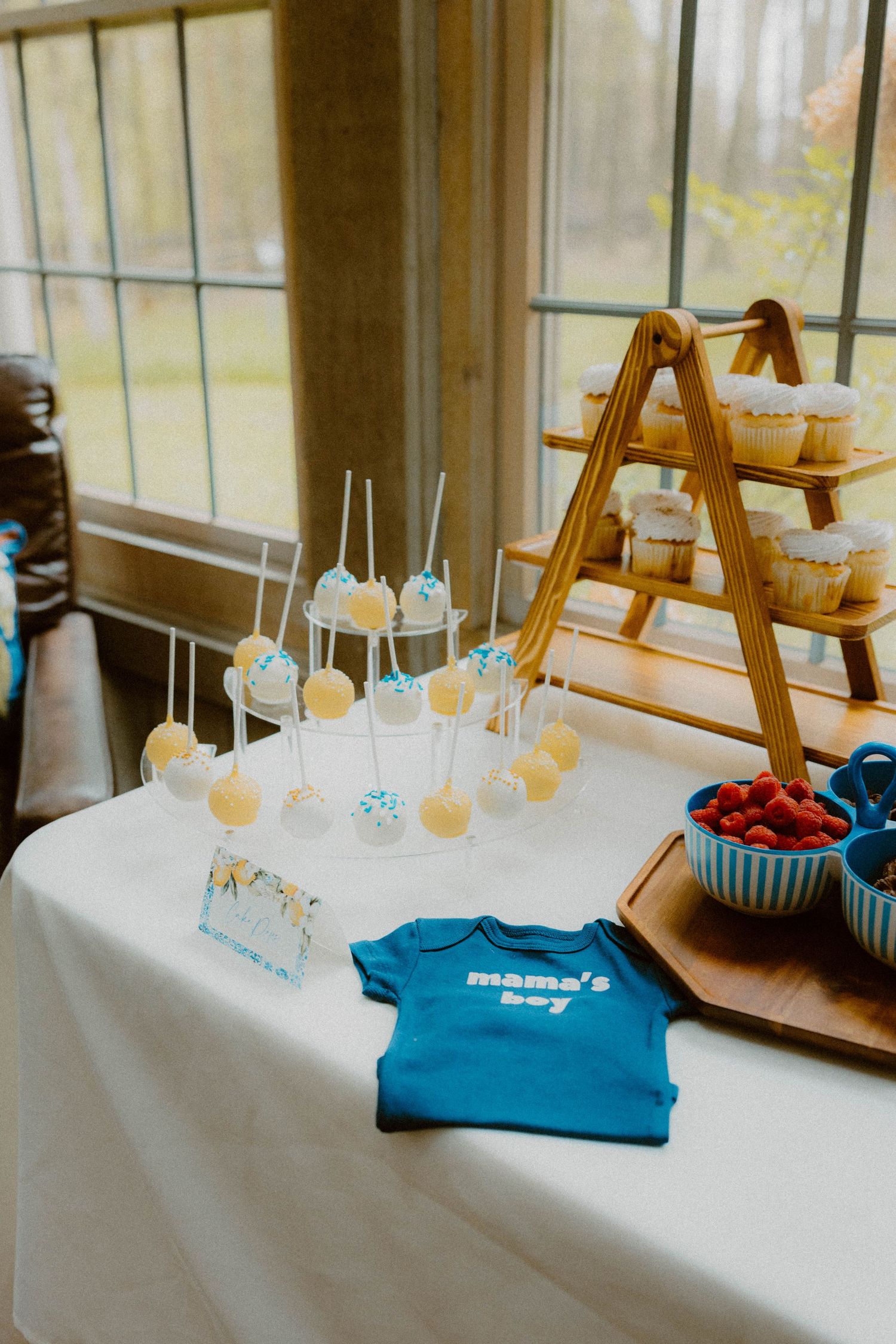 Baby shower dessert table with cake pops, cupcakes, and a blue mama's boy onesie on display.