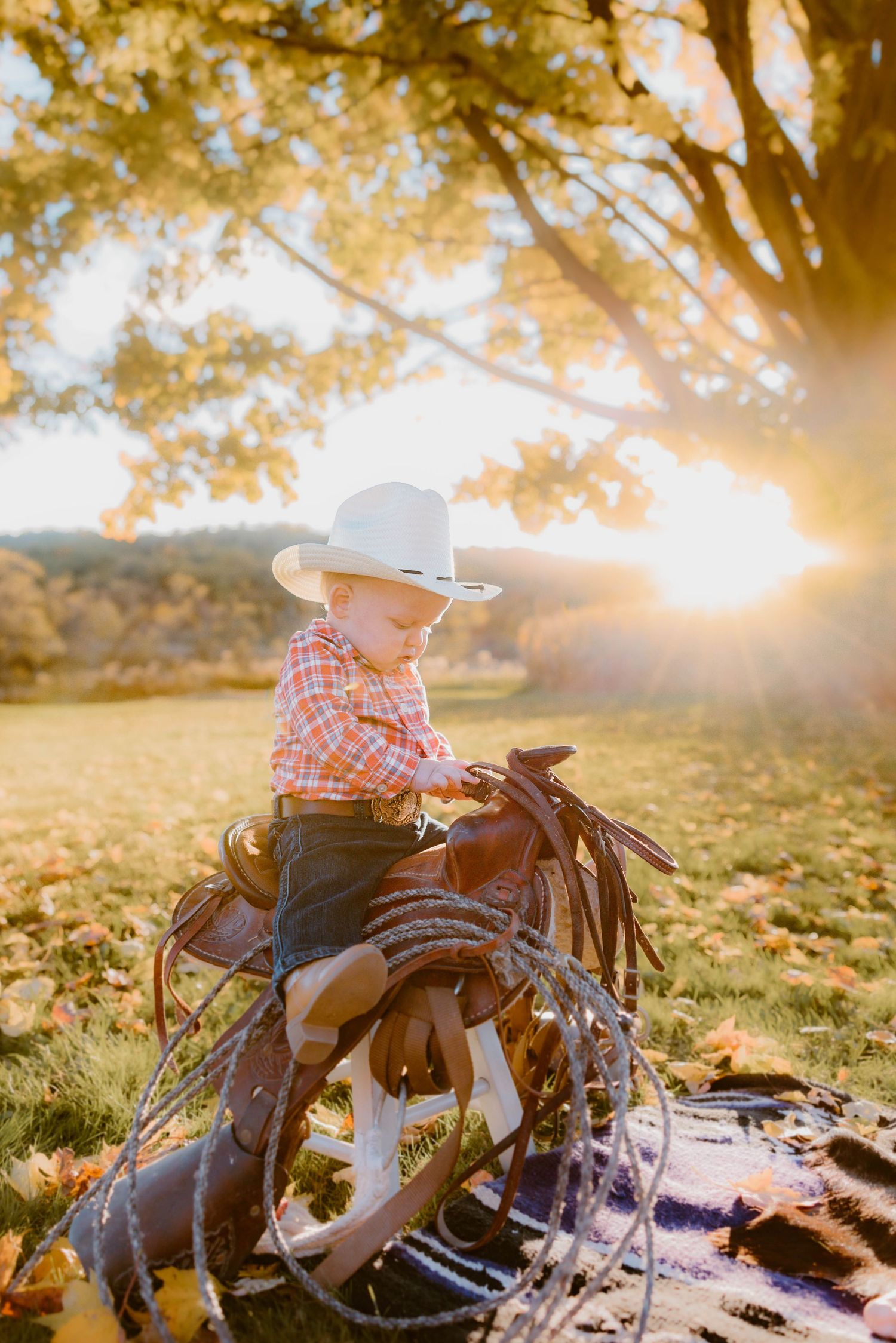 Adorable toddler wearing a cowboy hat sits on a saddle surrounded by golden autumn leaves at sunset.