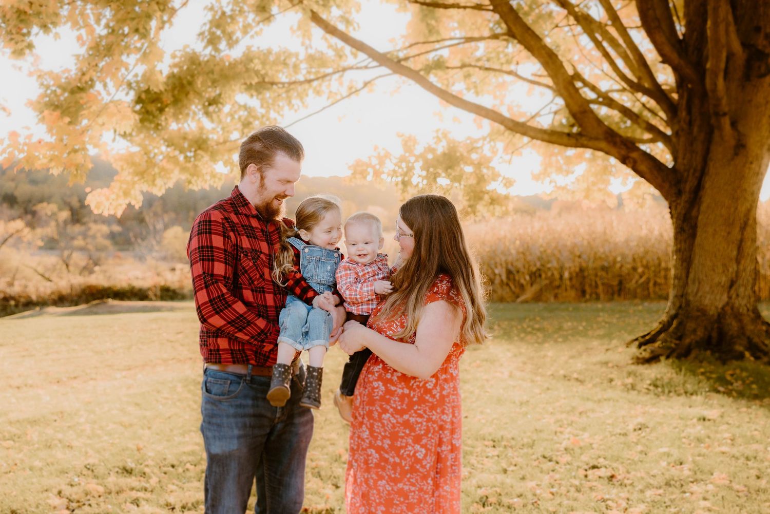 Family of three with parents smiling at their baby boy under a golden autumn tree outdoors.