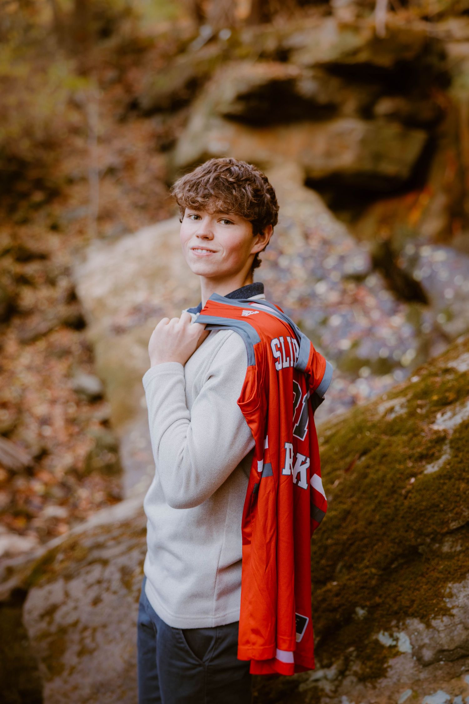 Smiling teen boy holding a red basketball jersey over his shoulder in a rocky outdoor setting.