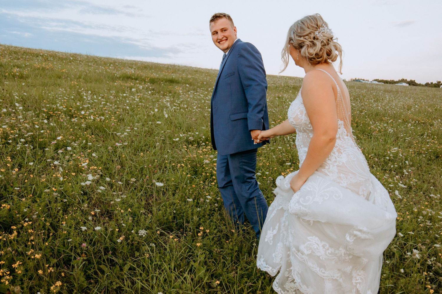 Smiling groom in blue suit leads bride through a wildflower meadow on their wedding day.