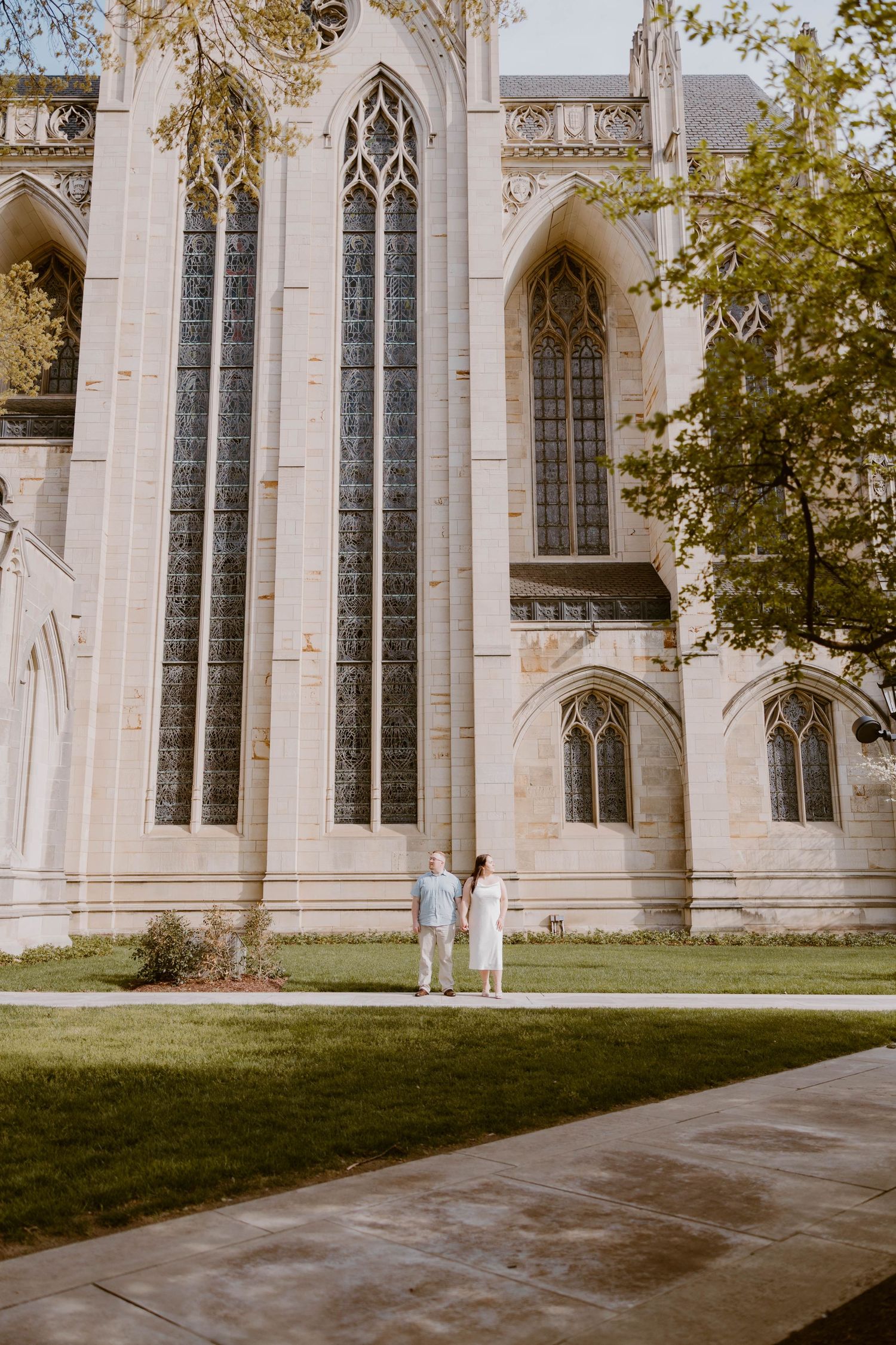 Engaged couple stands hand in hand on a lawn before a grand Gothic stone church with tall stained glass windows.