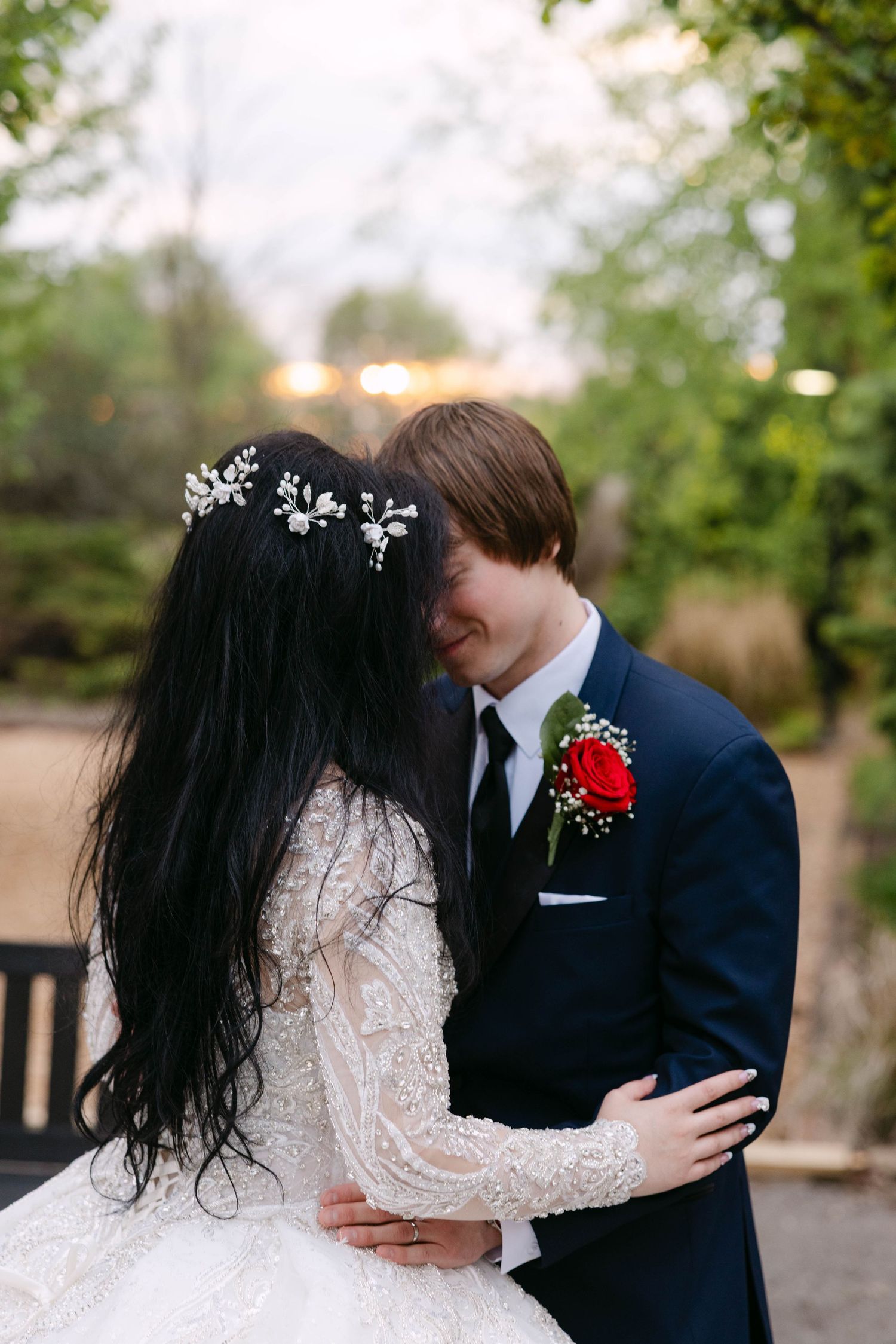 Bride and groom share an intimate moment, bride in long-sleeve lace gown with floral hair piece, groom in navy suit.