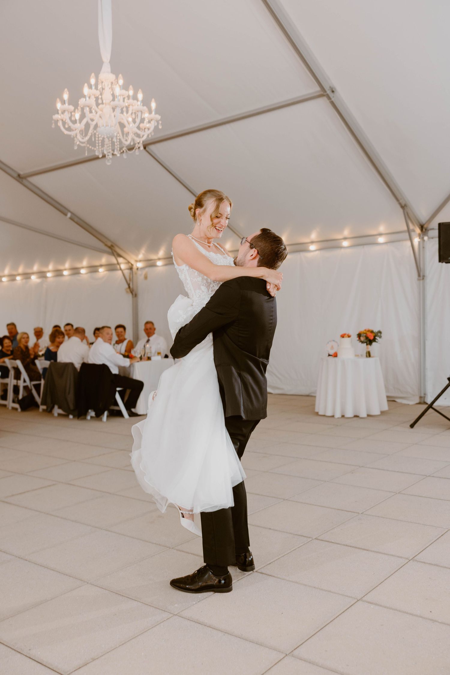 Groom lifting bride during first dance under a chandelier in a white tent reception.